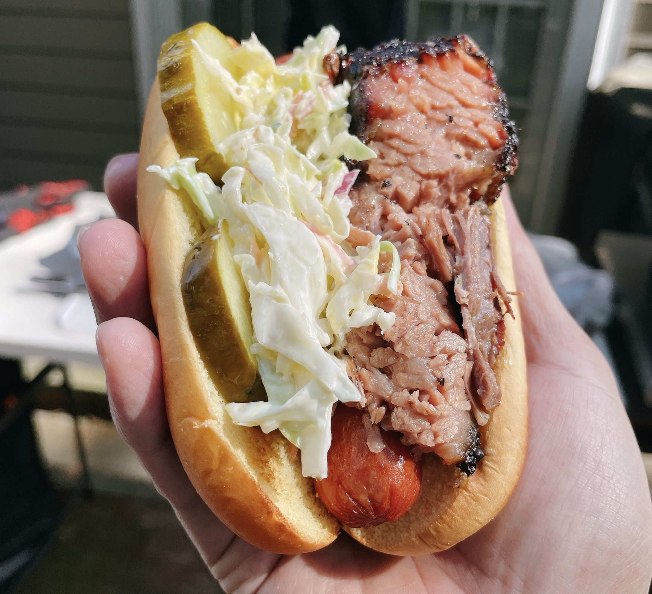 A hand holds a hot dog in a bun topped with sliced pickles, coleslaw, and a thick piece of smoked brisket.