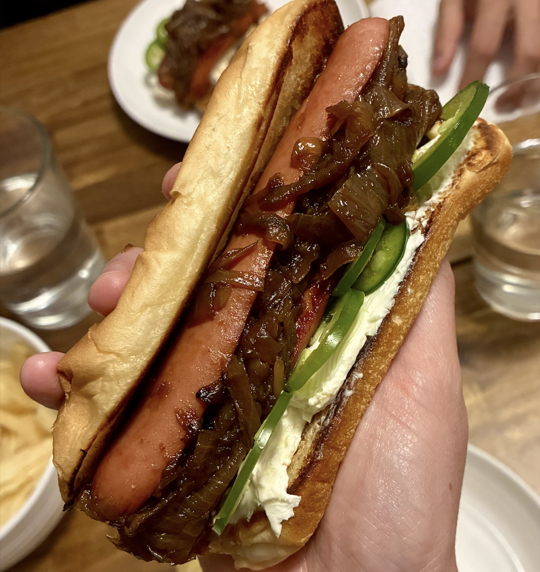 A hand holds a hot dog topped with sautƩed onions, sliced jalapeƱos, and cream cheese in a toasted bun. Plates of food and glasses of water are on the wooden table in the background.
