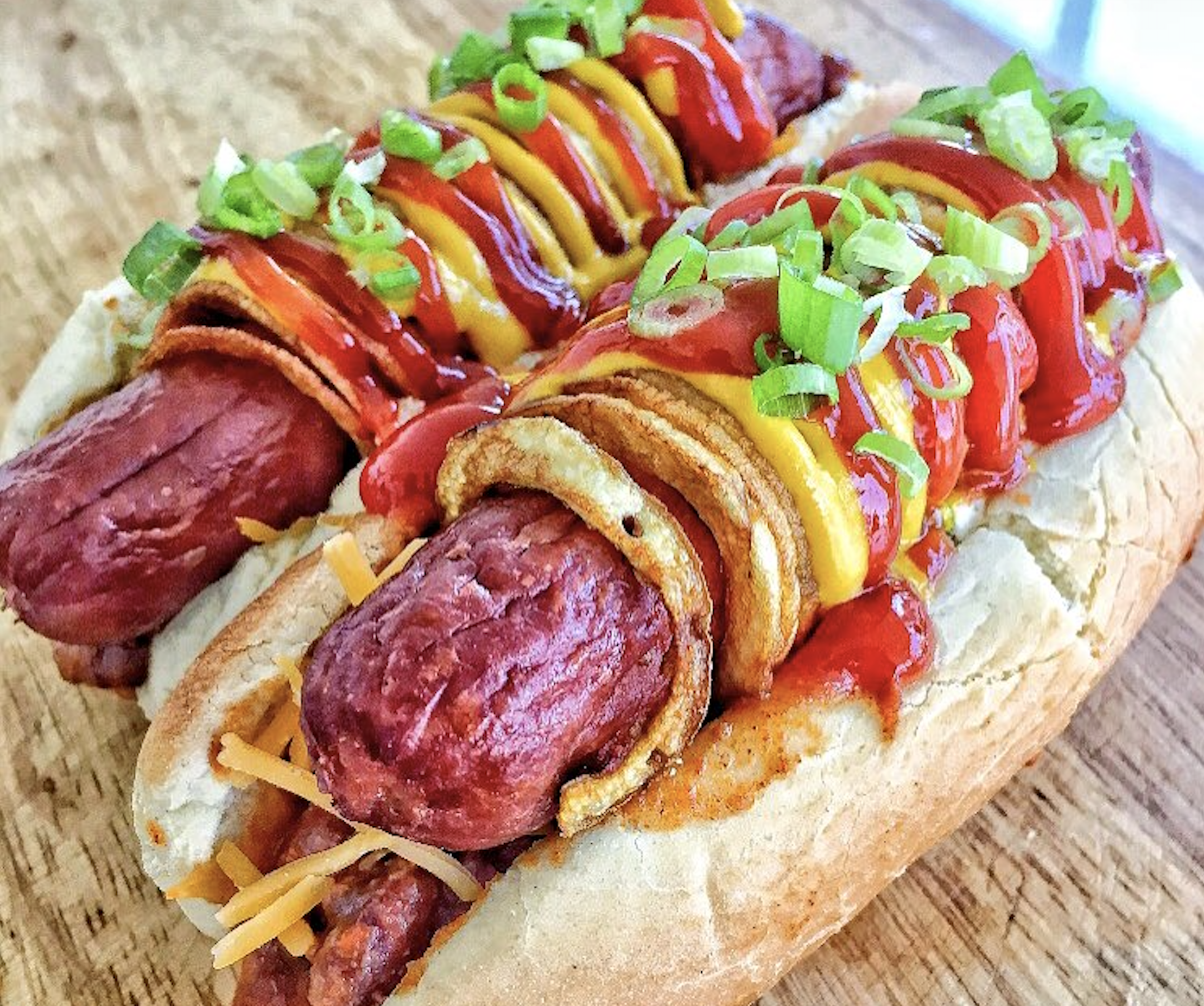 Two grilled hot dogs in buns topped with crispy onion rings, shredded cheese, ketchup, mustard, and chopped green onions, served on a wooden surface.