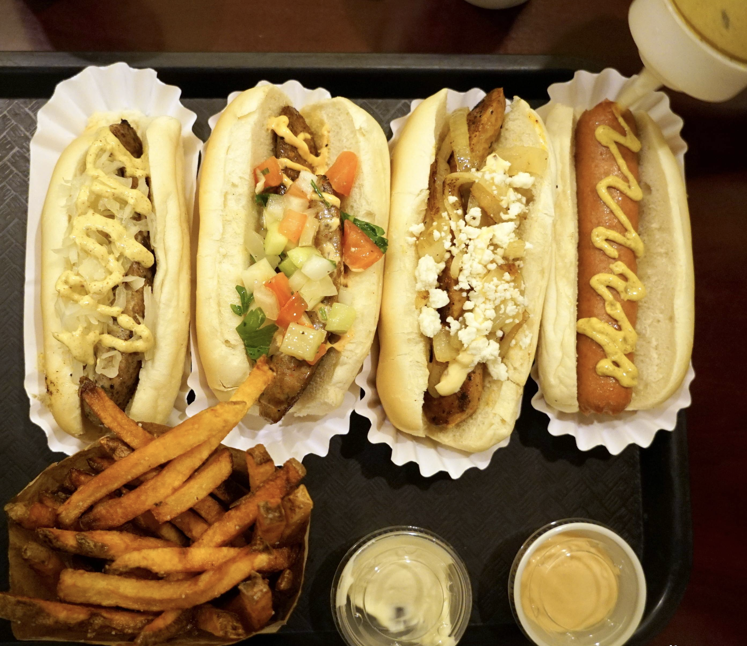 Four gourmet hot dogs with various toppings are lined up on a tray, accompanied by a serving of French fries and two dipping sauces in small containers.