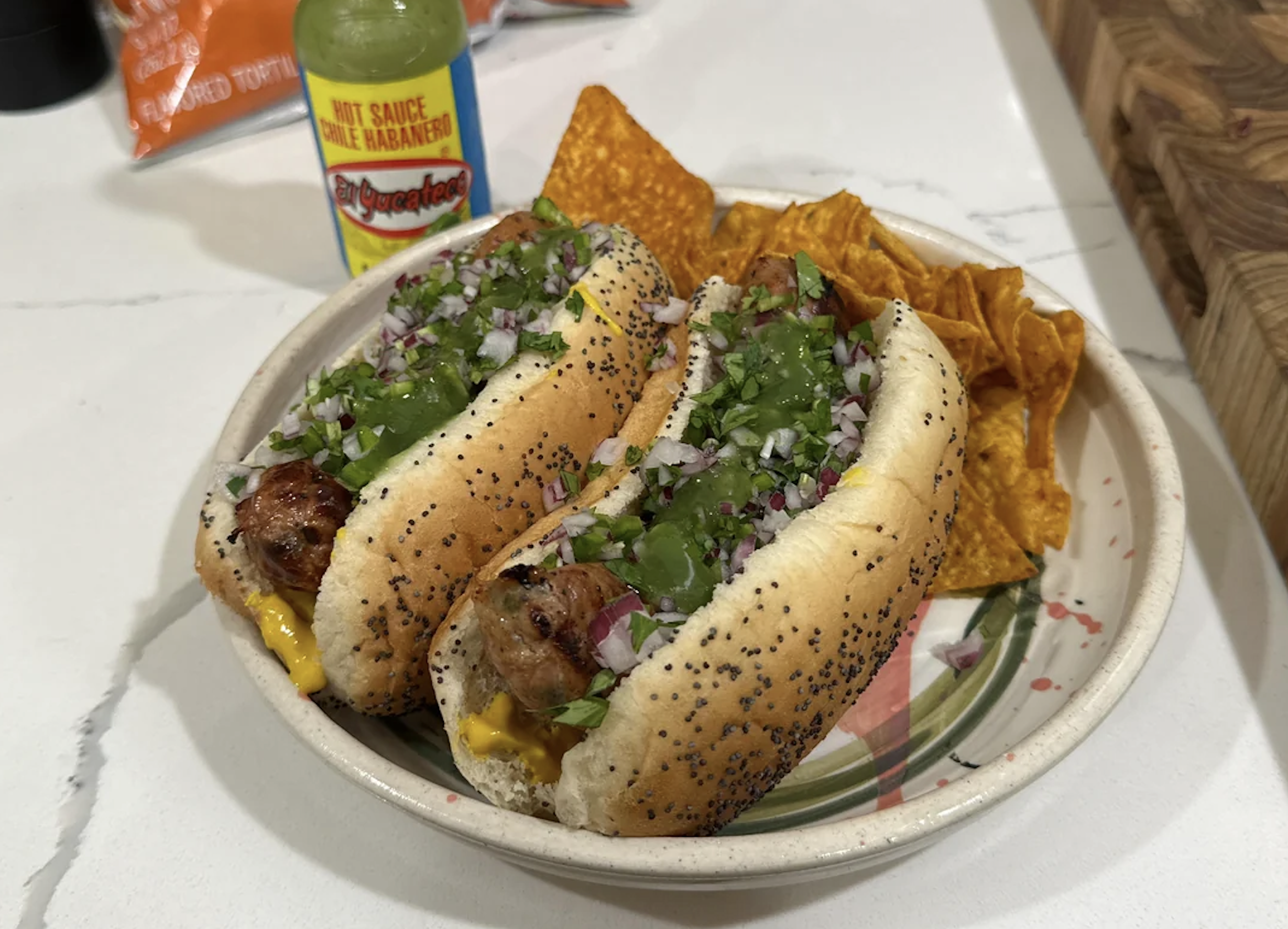 Two poppy seed hot dogs topped with relish, onions, and cilantro on a plate with nacho cheese chips. A bottle of green hot sauce is nearby on a white countertop.