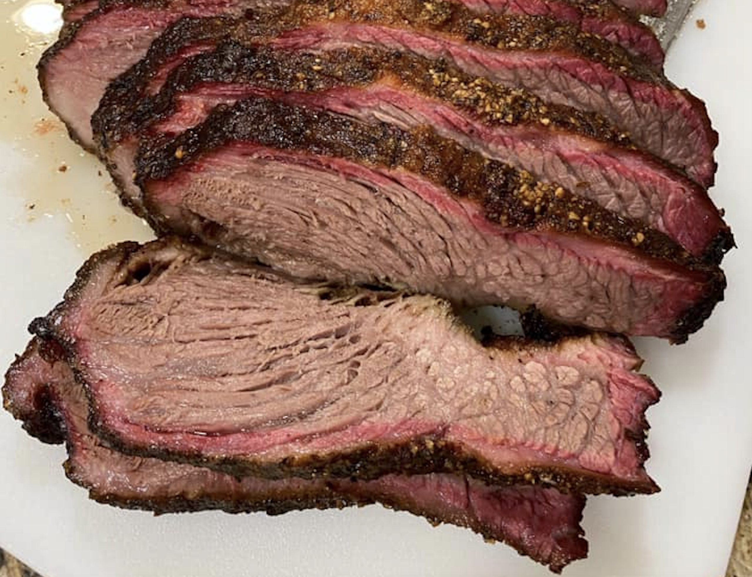 Sliced smoked brisket on a white cutting board, showing a dark seasoned crust, a pink smoke ring, and tender, juicy meat inside.