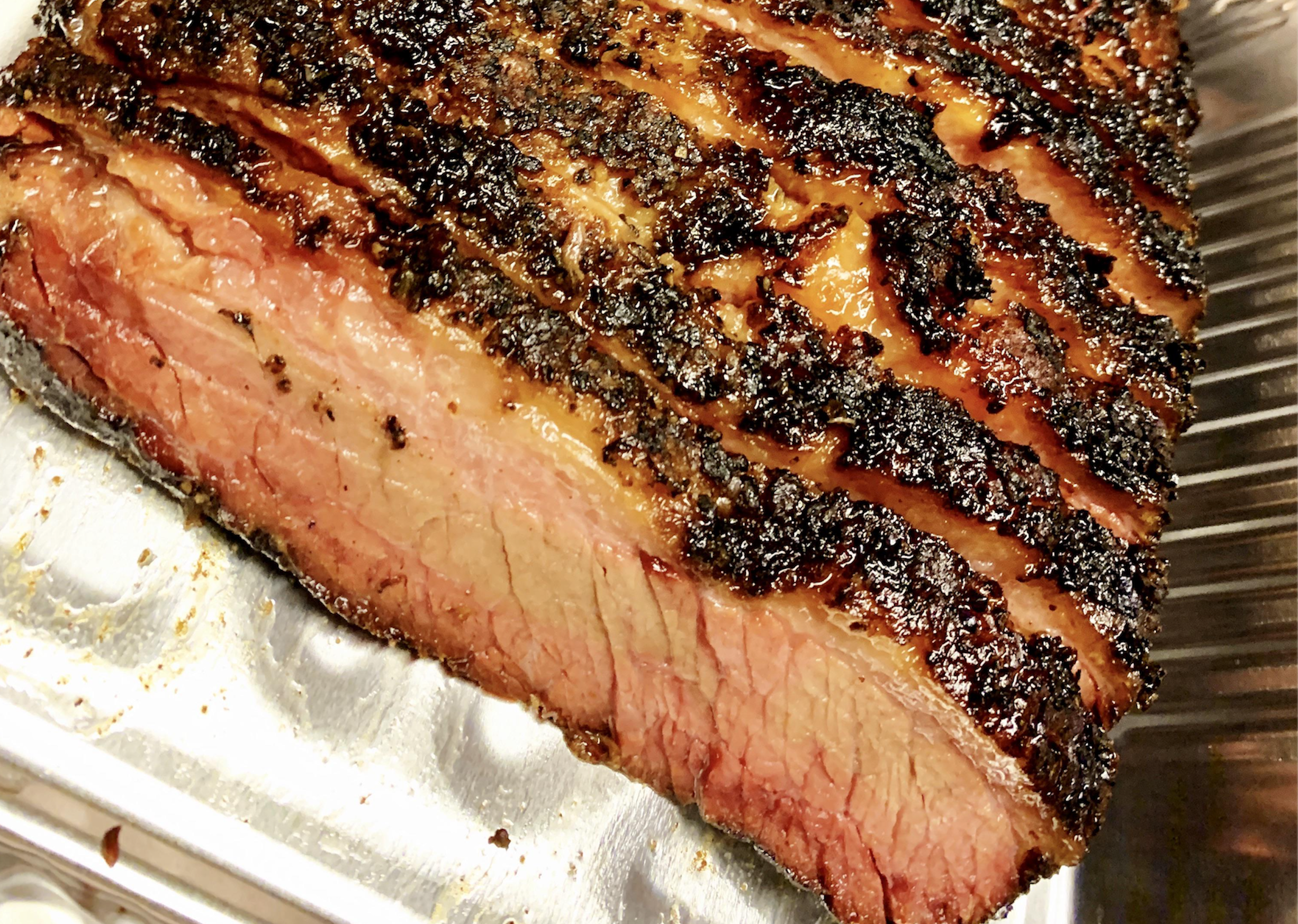 A close-up of sliced smoked brisket with a charred, seasoned crust and a pink smoke ring, resting in a metal tray.