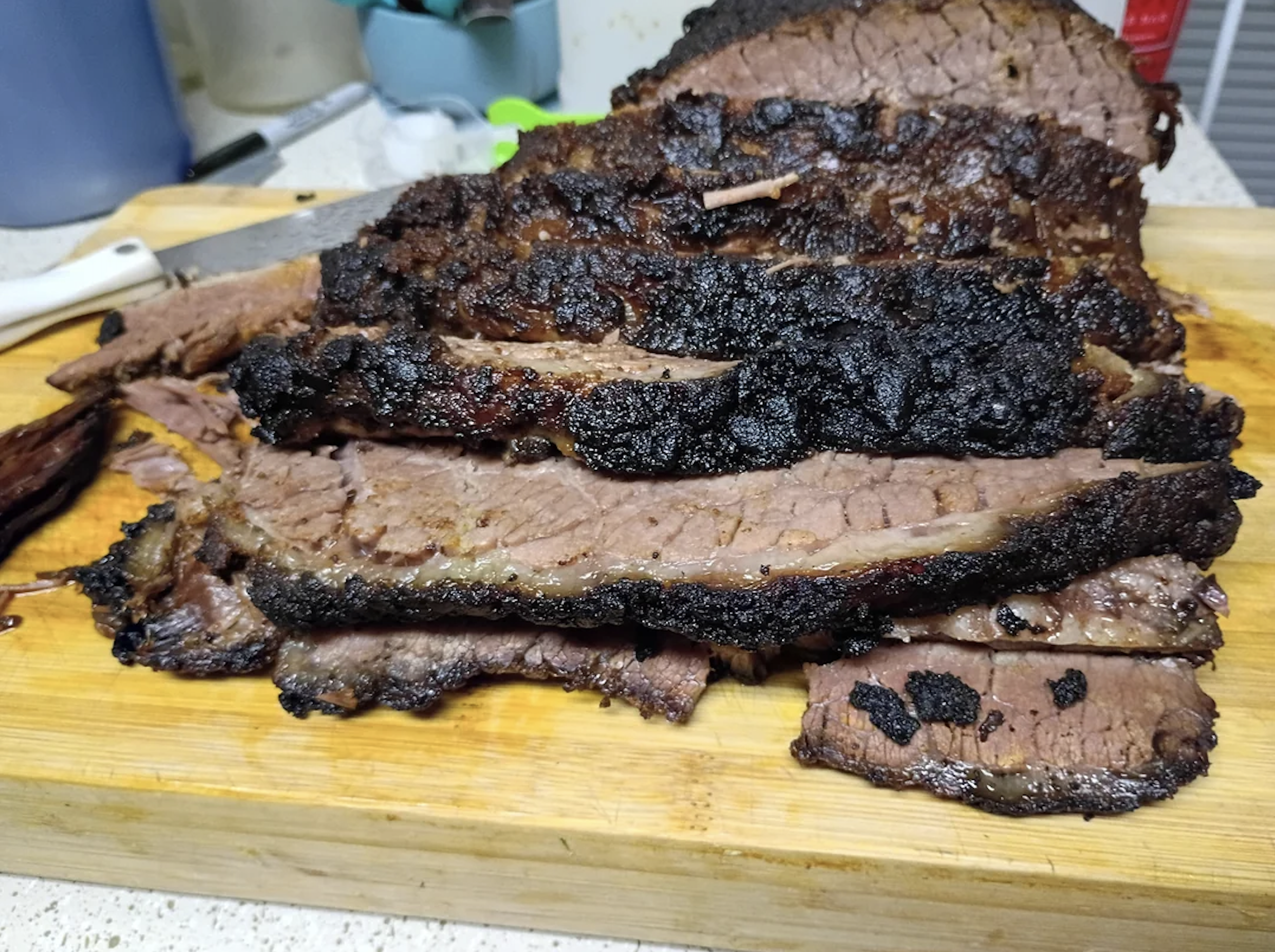 Sliced smoked brisket with a dark, crispy bark stacked on a wooden cutting board, showing juicy, tender meat inside. Kitchen utensils and containers are visible in the background.