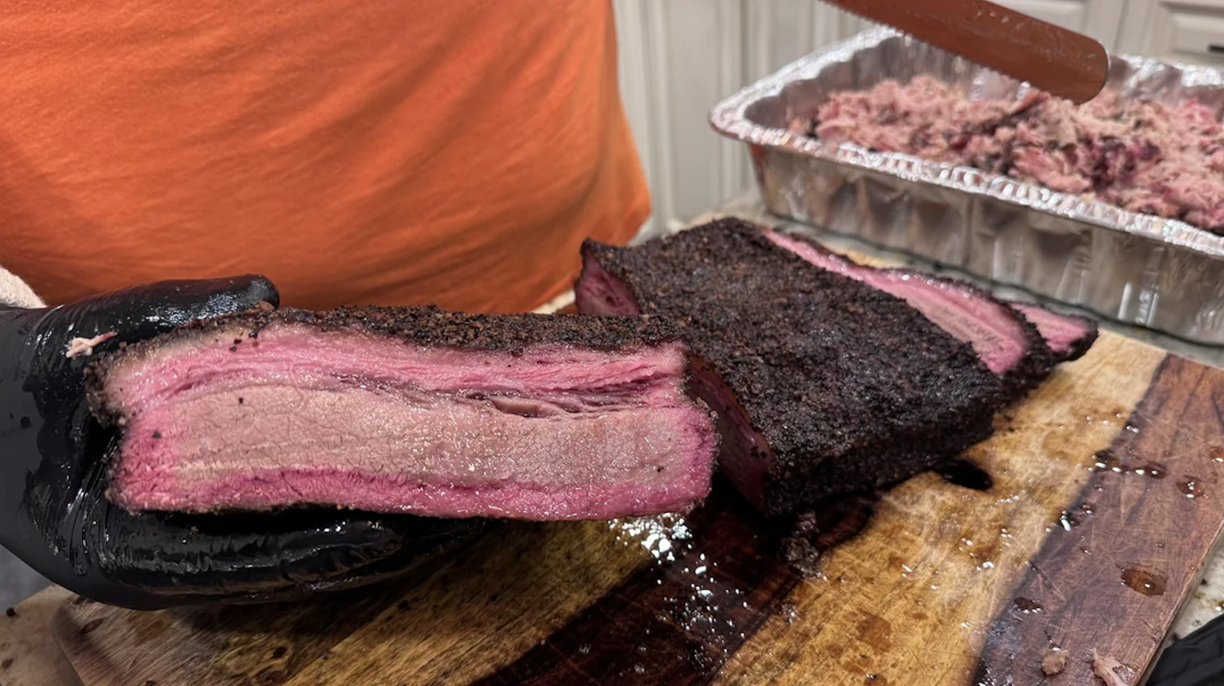 A person in an orange shirt slices a juicy, smoked brisket on a wooden cutting board. A pan of shredded meat is visible in the background.