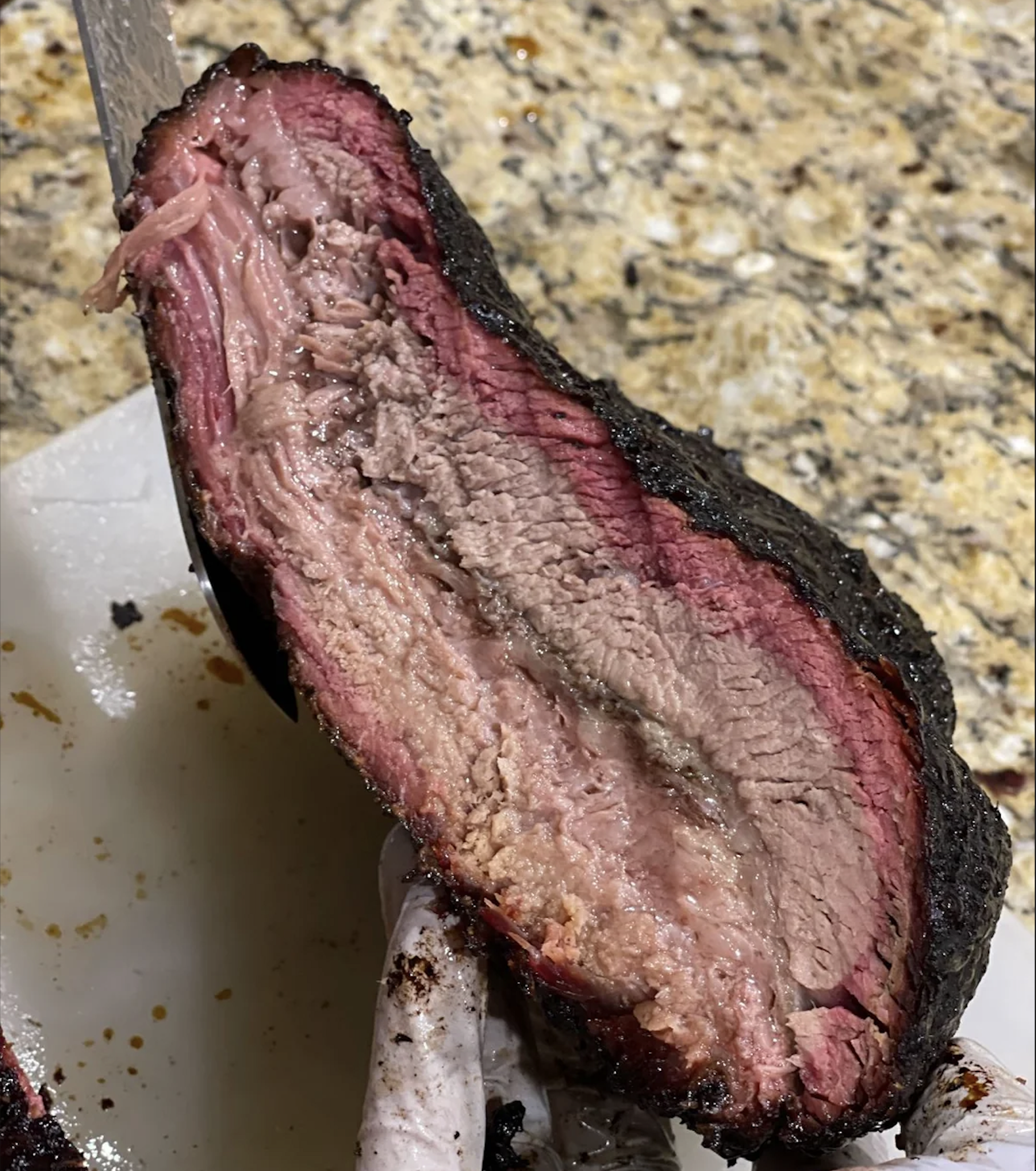 A close-up of a sliced smoked brisket showing a dark bark, a pink smoke ring, and juicy, tender meat. The brisket is held over a cutting board on a granite countertop.