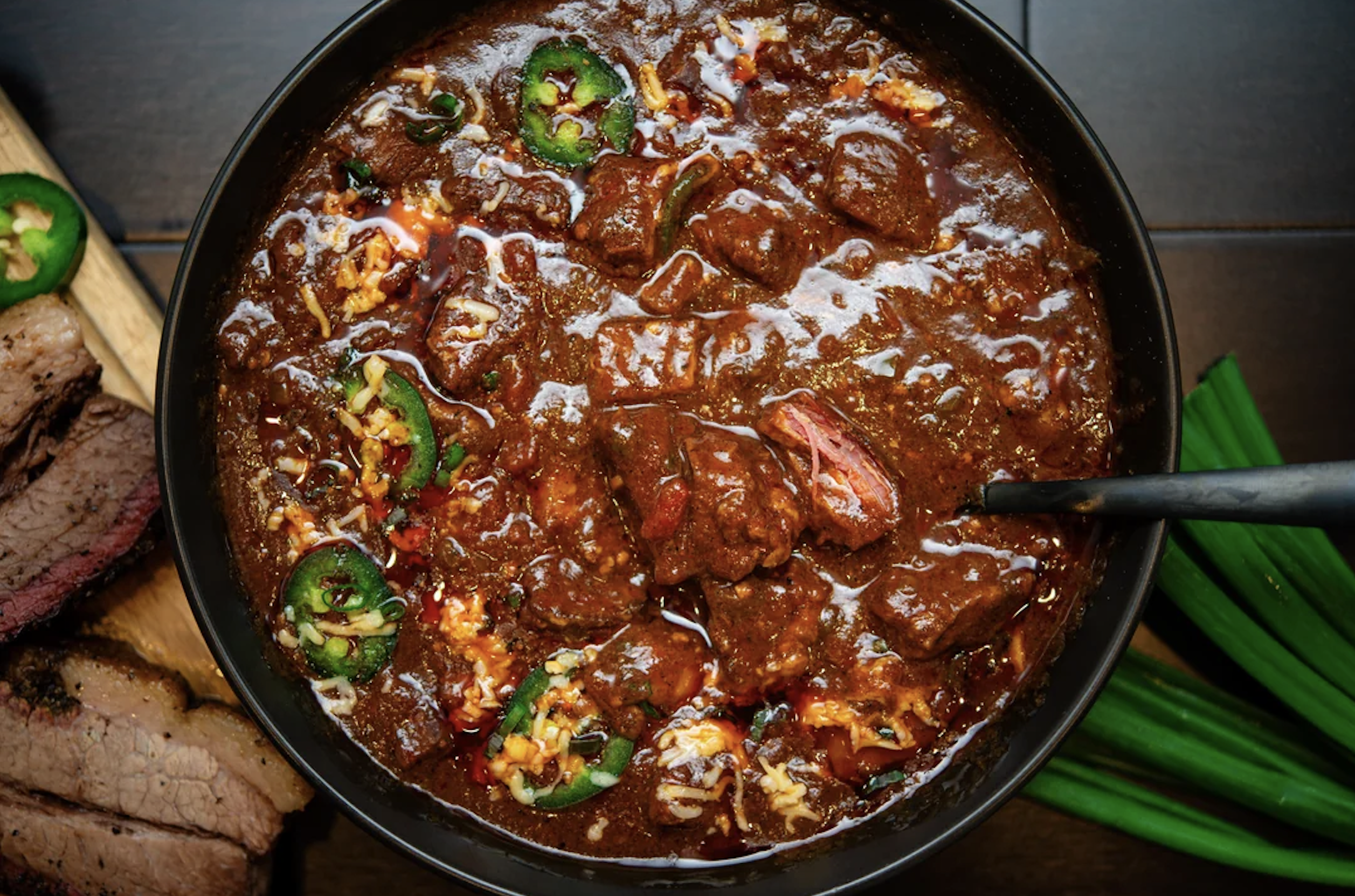 A bowl of spicy beef chili topped with jalapeño slices and cheese, next to sliced brisket and fresh green onions on a dark table.