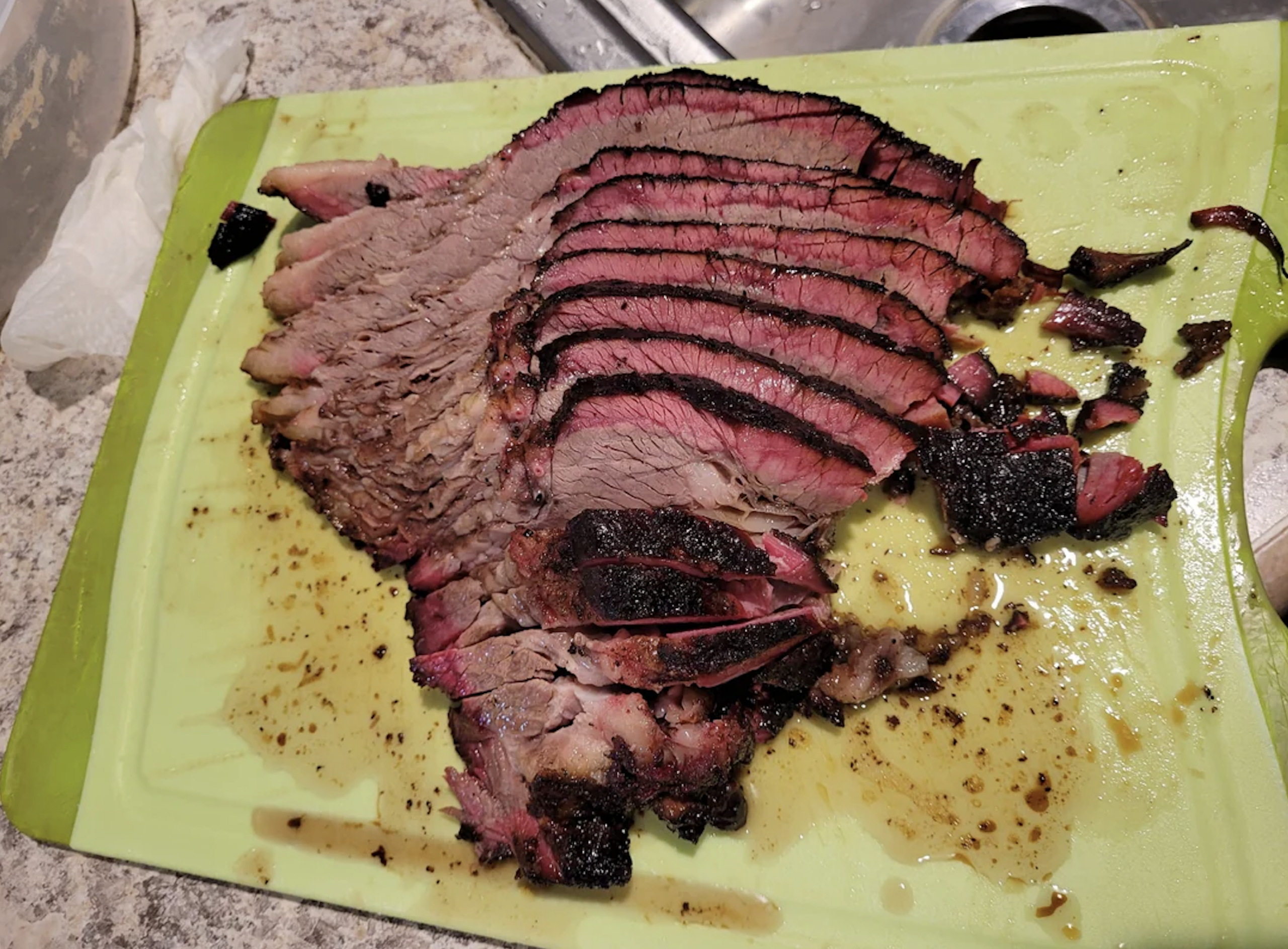 Slices of smoked brisket with a dark bark and pink smoke ring are arranged on a green cutting board, with juices pooling around the meat on a kitchen counter.