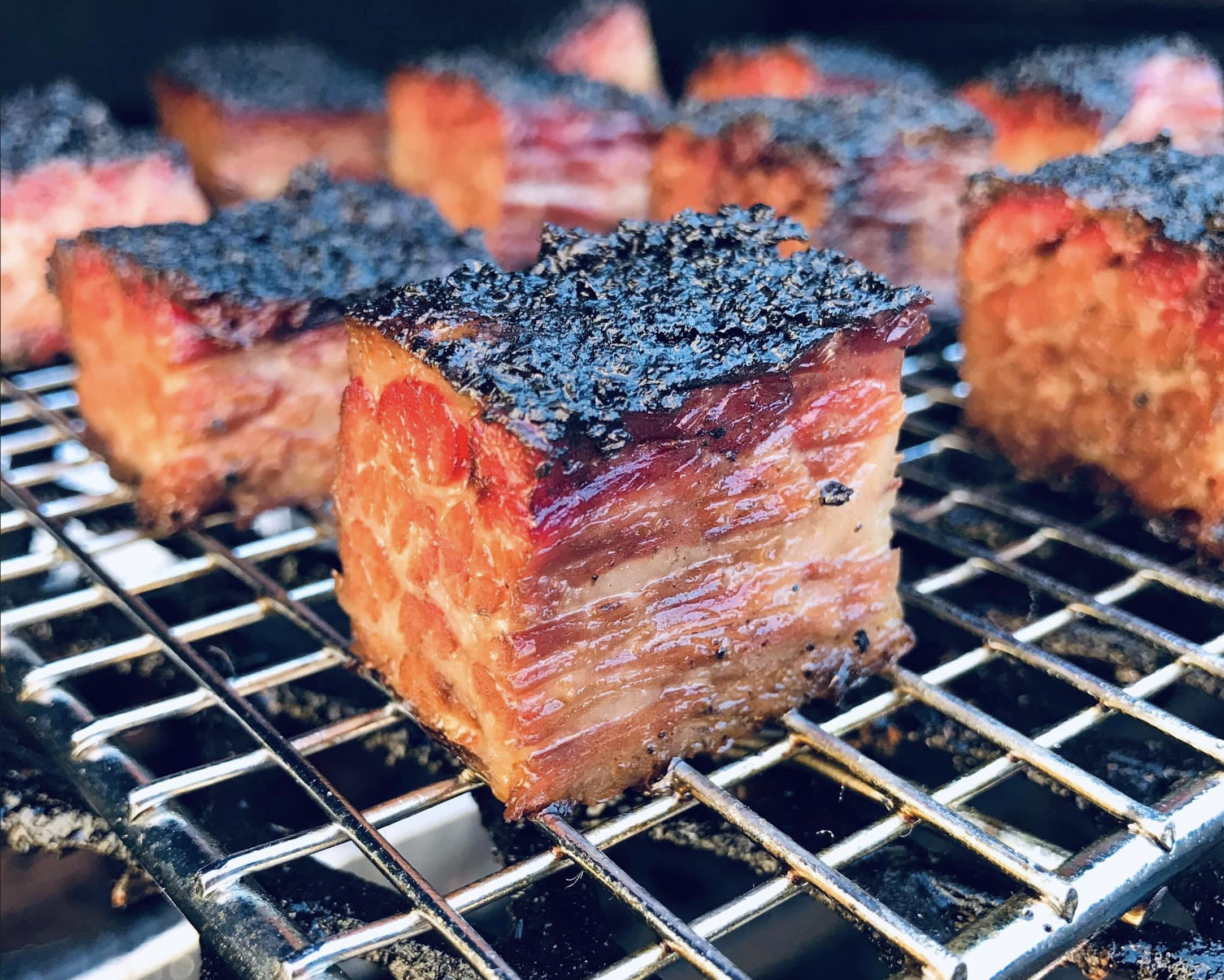Close-up of several pieces of smoked brisket burnt ends on a metal grill, showing a crispy, dark bark on top and juicy, marbled meat layers beneath.