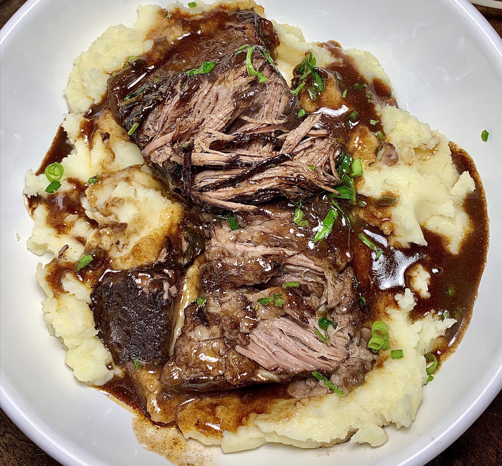 Shredded pot roast with rich brown gravy served over creamy mashed potatoes, garnished with chopped green onions in a white bowl.