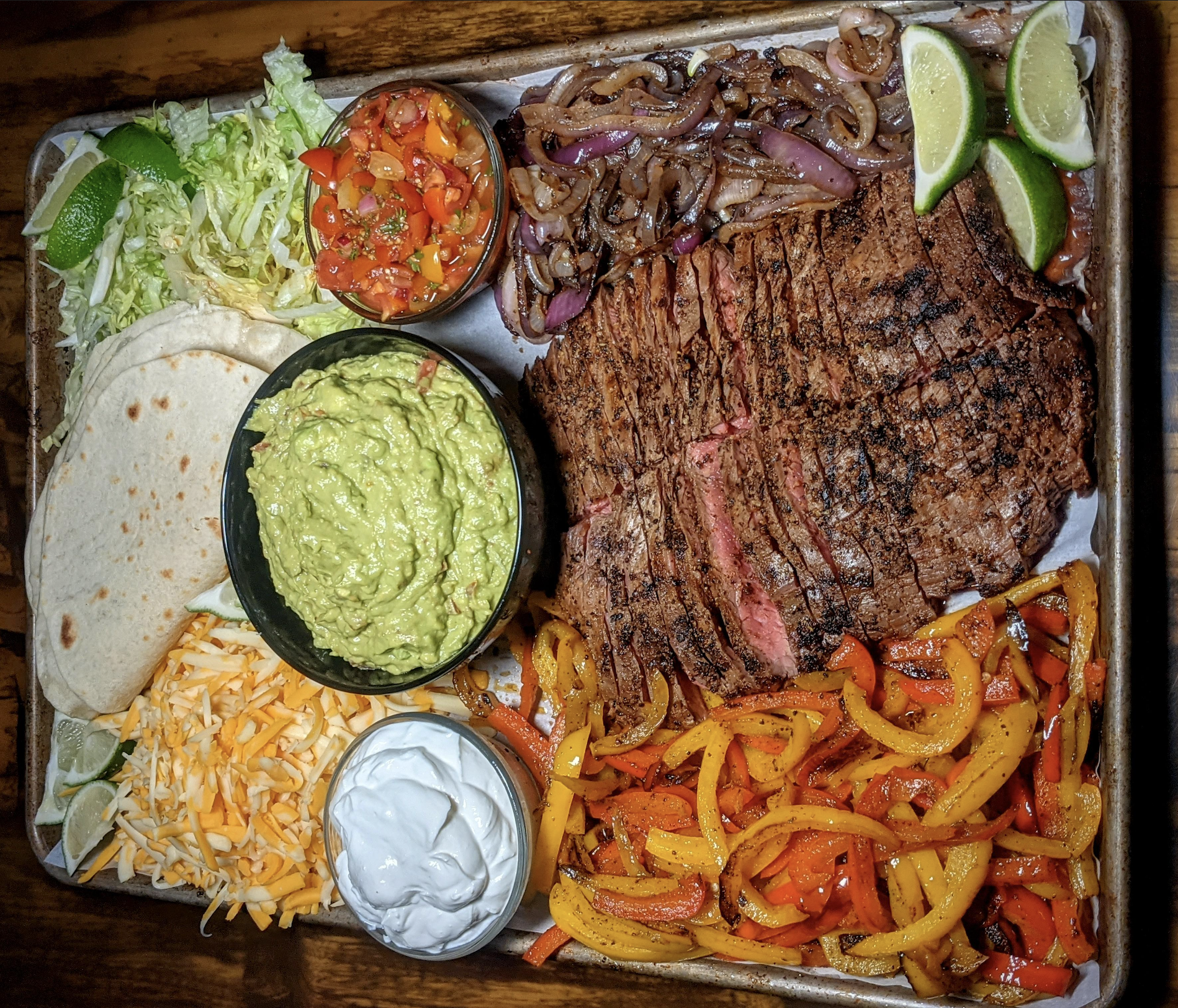 A tray with grilled steak, sautéed onions, bell peppers, shredded lettuce, cheese, guacamole, salsa, sour cream, tortillas, and lime wedges, arranged in sections for a fajita meal.