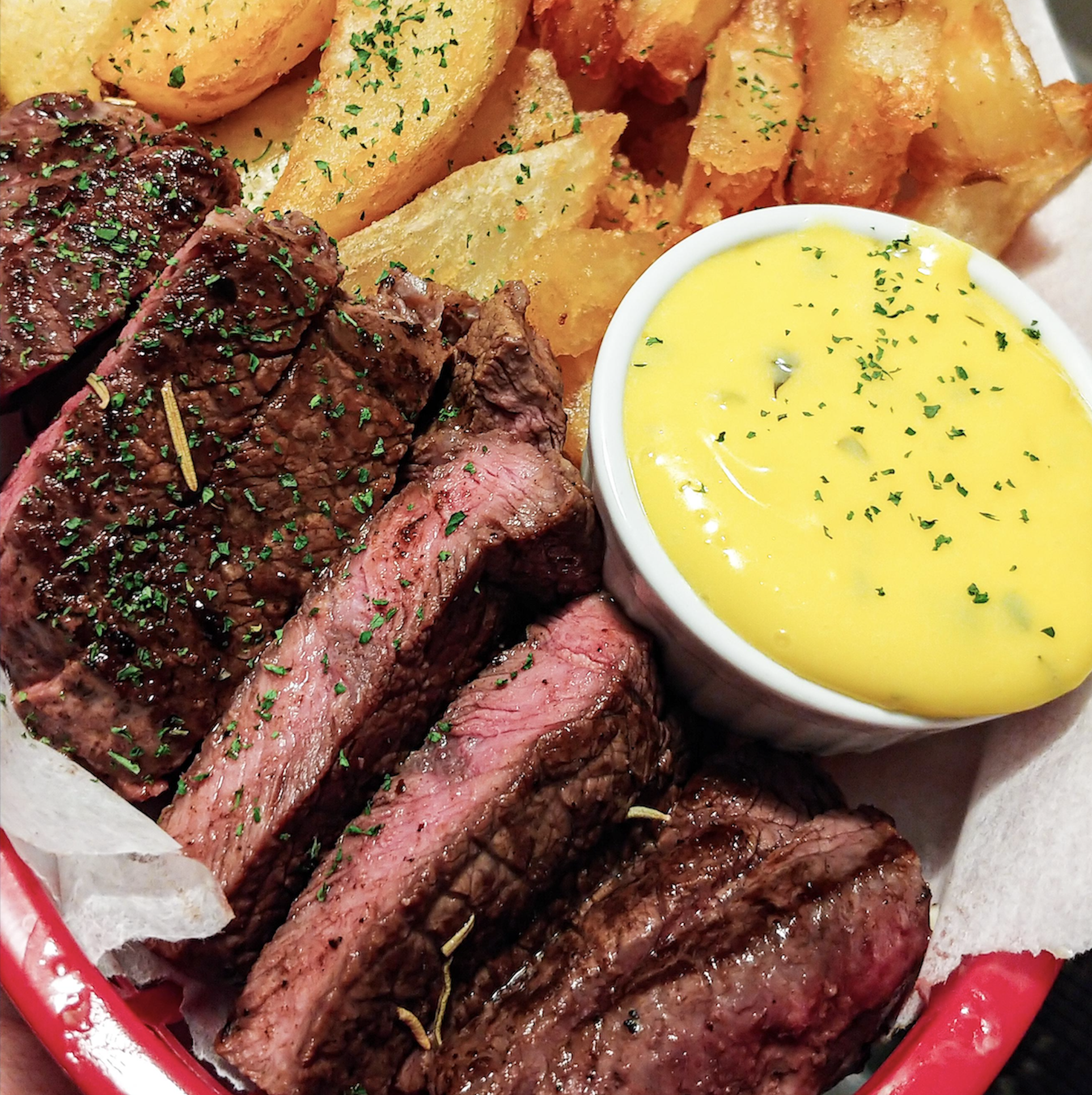 Sliced grilled steak with herbs, crispy potato wedges, and a small bowl of yellow sauce, garnished with chopped parsley, served on a white plate.