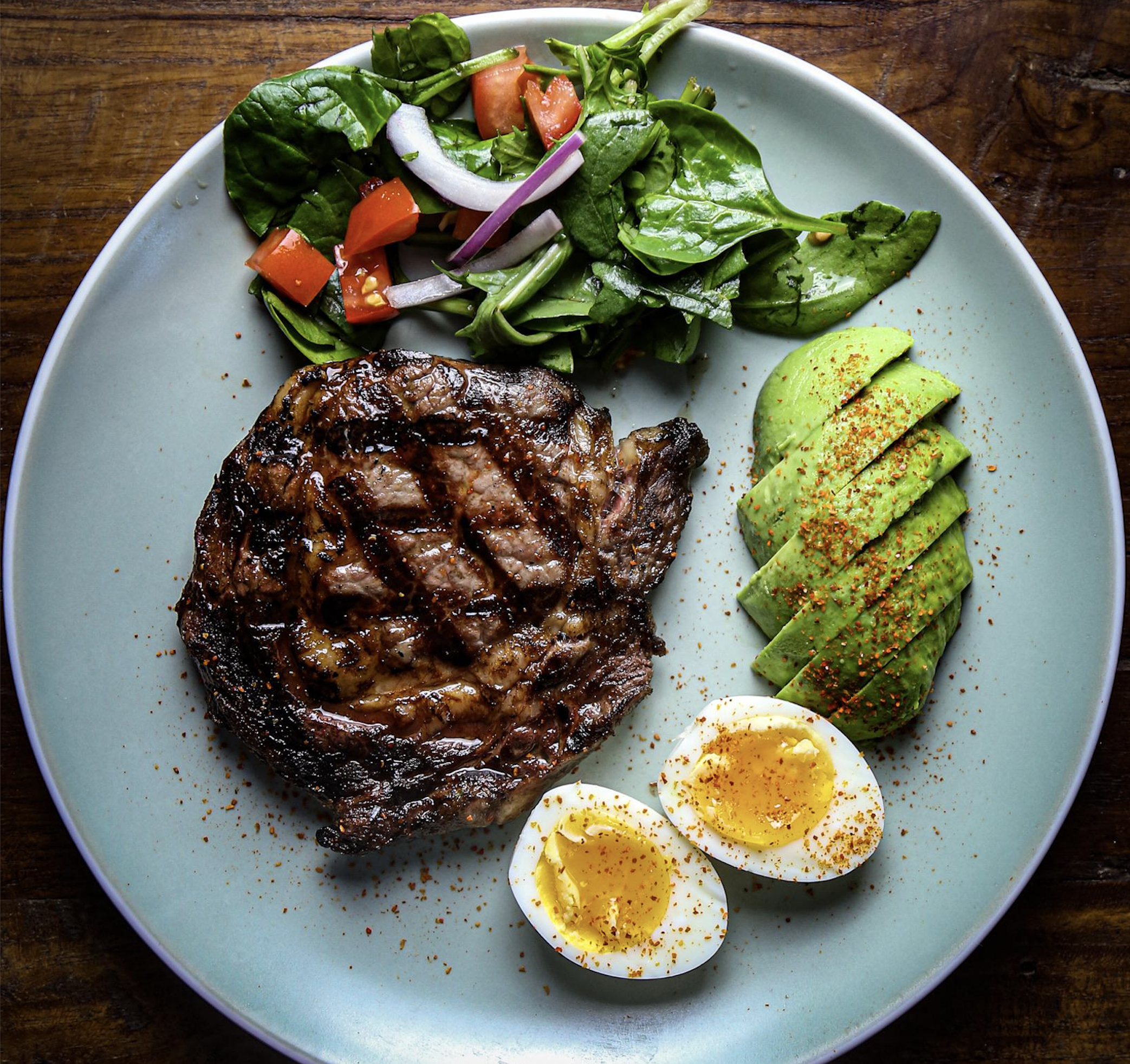A plate with grilled steak, halved soft-boiled egg, sliced avocado, and a spinach salad with tomato and red onion. The food is sprinkled with seasoning and served on a light blue plate atop a wooden table.