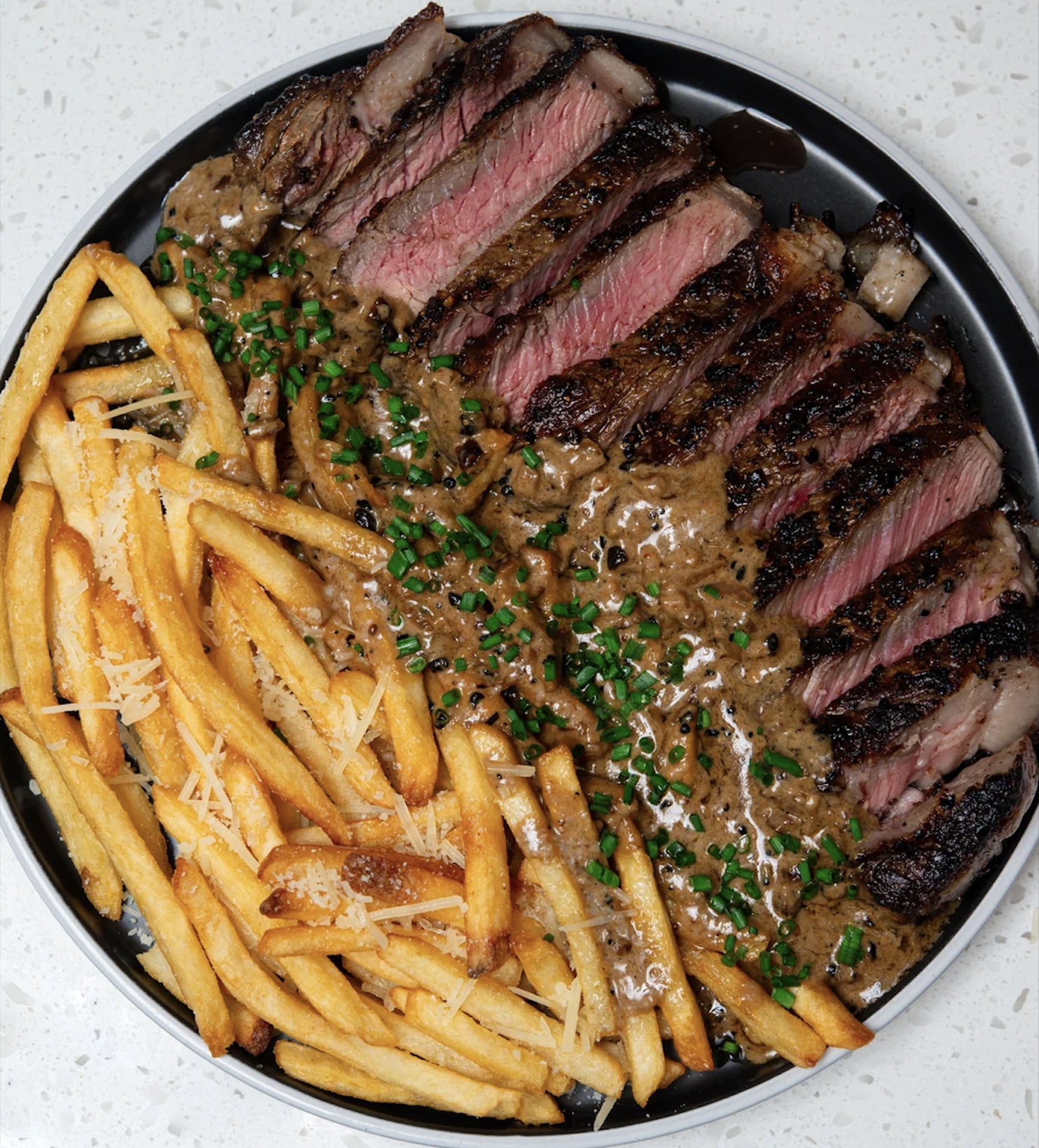 Sliced medium-rare steak with brown sauce and chopped chives, served with golden fries topped with grated cheese, on a black plate over a white surface.
