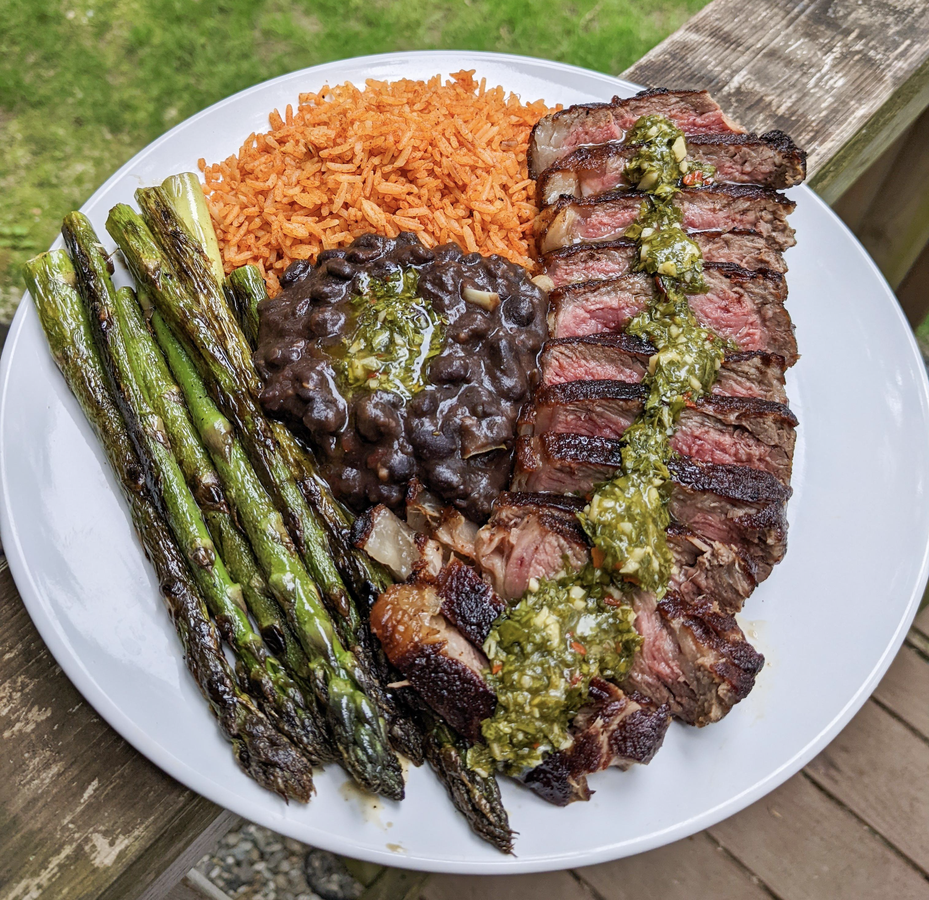 A plate with grilled asparagus, Mexican rice, black beans, and sliced steak topped with green sauce, placed on a wooden railing outdoors with grass in the background.