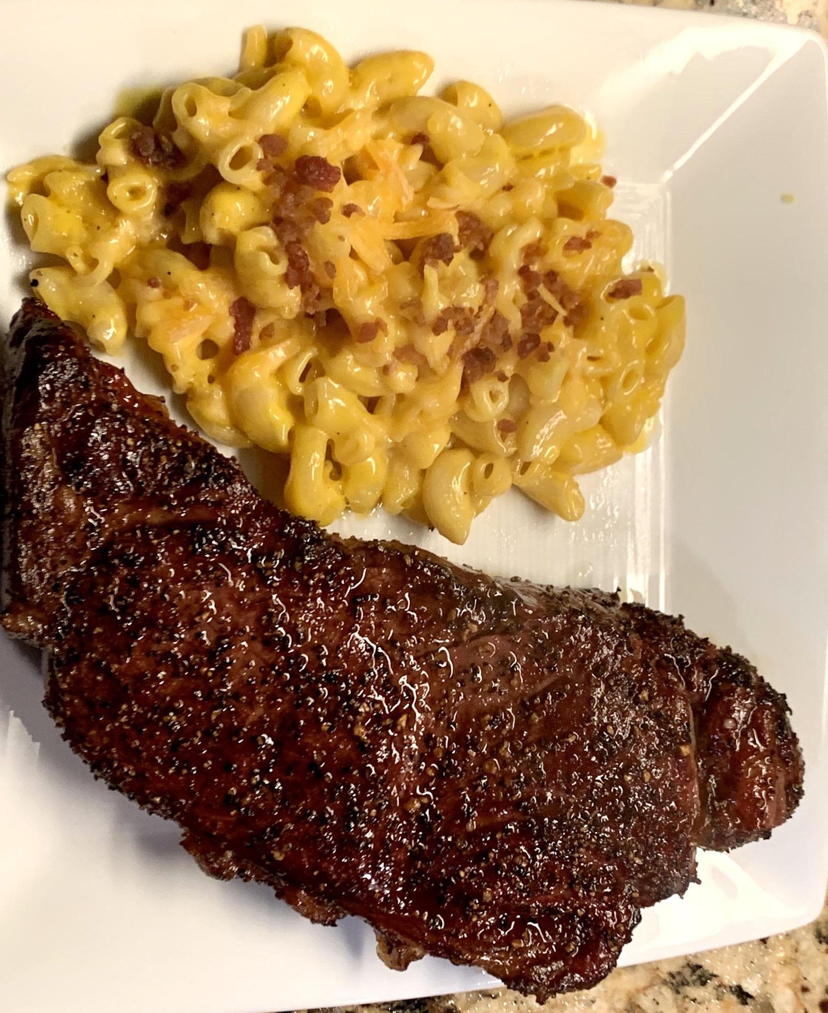 A grilled steak with a peppered crust is served next to creamy macaroni and cheese with bits of bacon on a white plate.