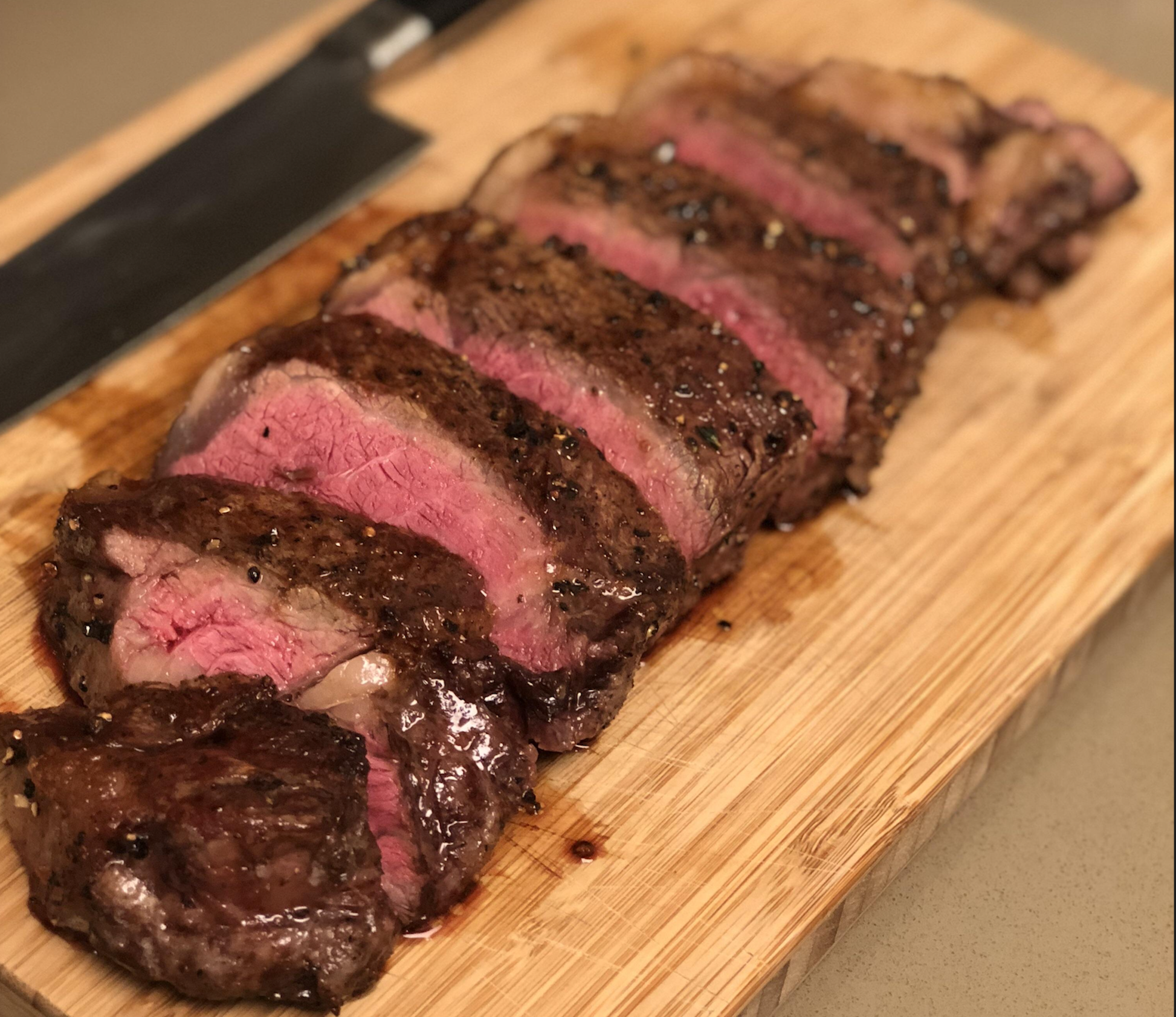 Sliced medium-rare steak with a seared crust is arranged on a wooden cutting board, with a large knife visible in the background.