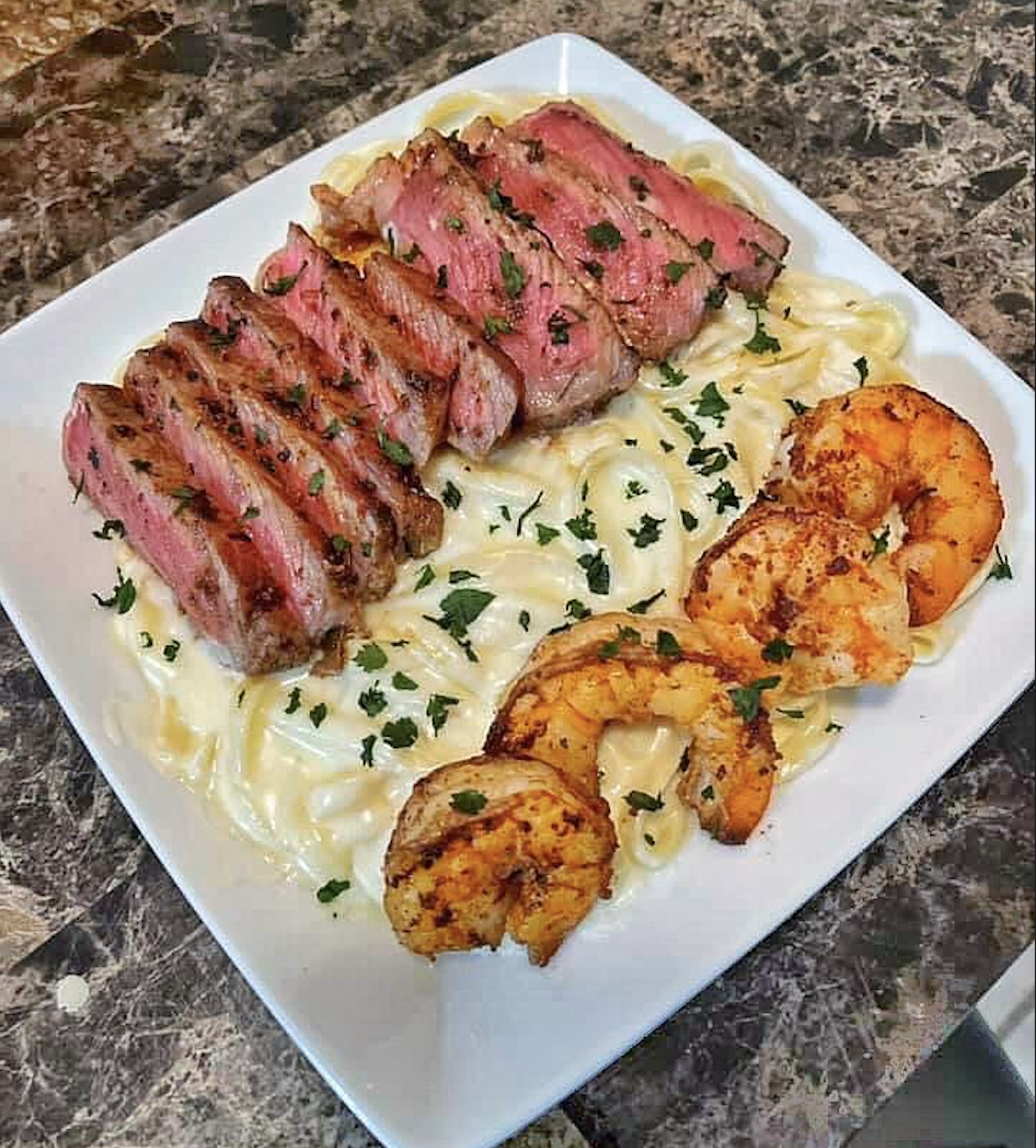 A square white plate with fettuccine Alfredo, topped with sliced steak and three grilled shrimp, garnished with chopped parsley, on a dark marble surface.