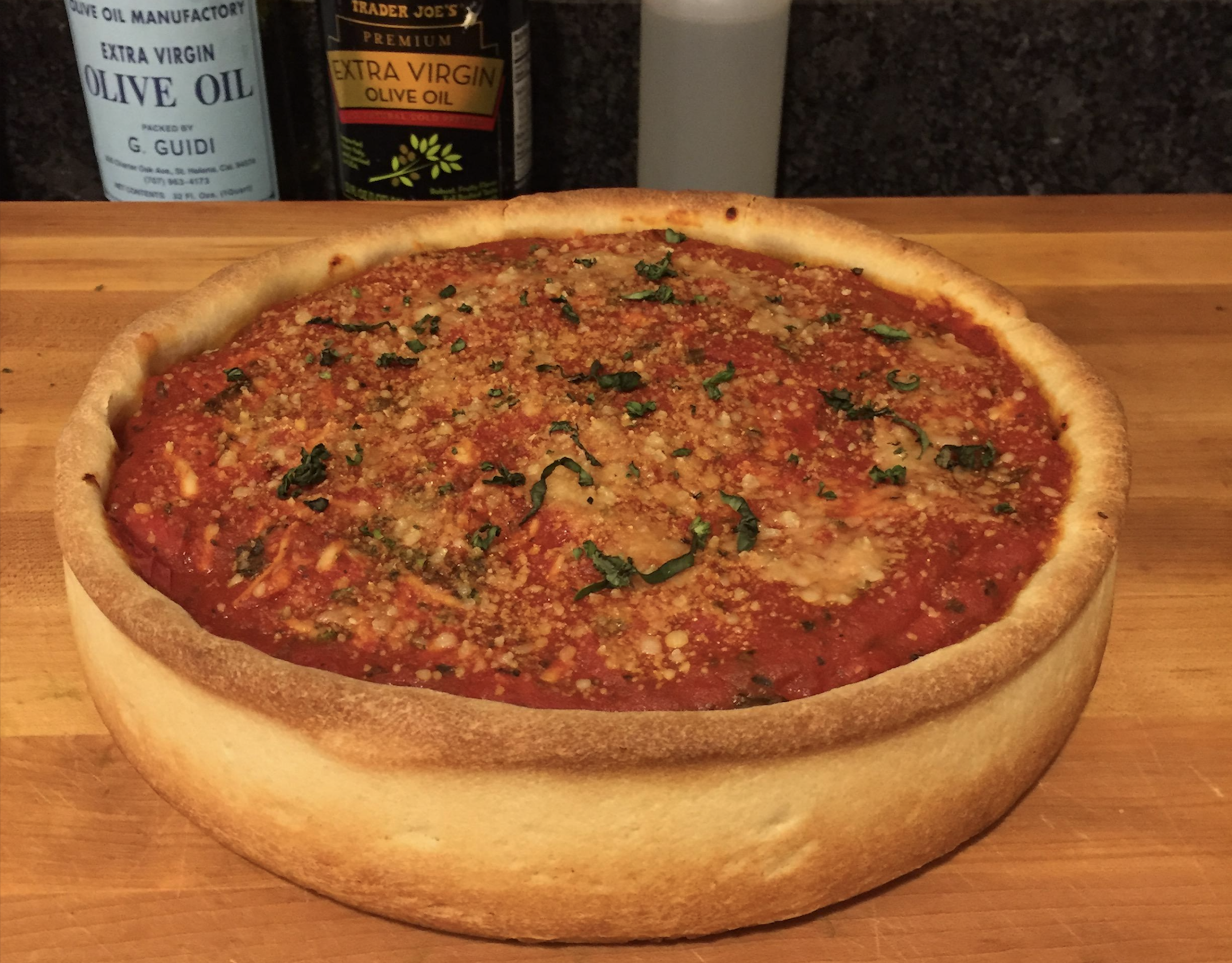A whole Chicago-style deep-dish pizza with thick golden crust, topped with tomato sauce, grated cheese, and fresh basil, sits on a wooden counter with bottles of olive oil in the background.