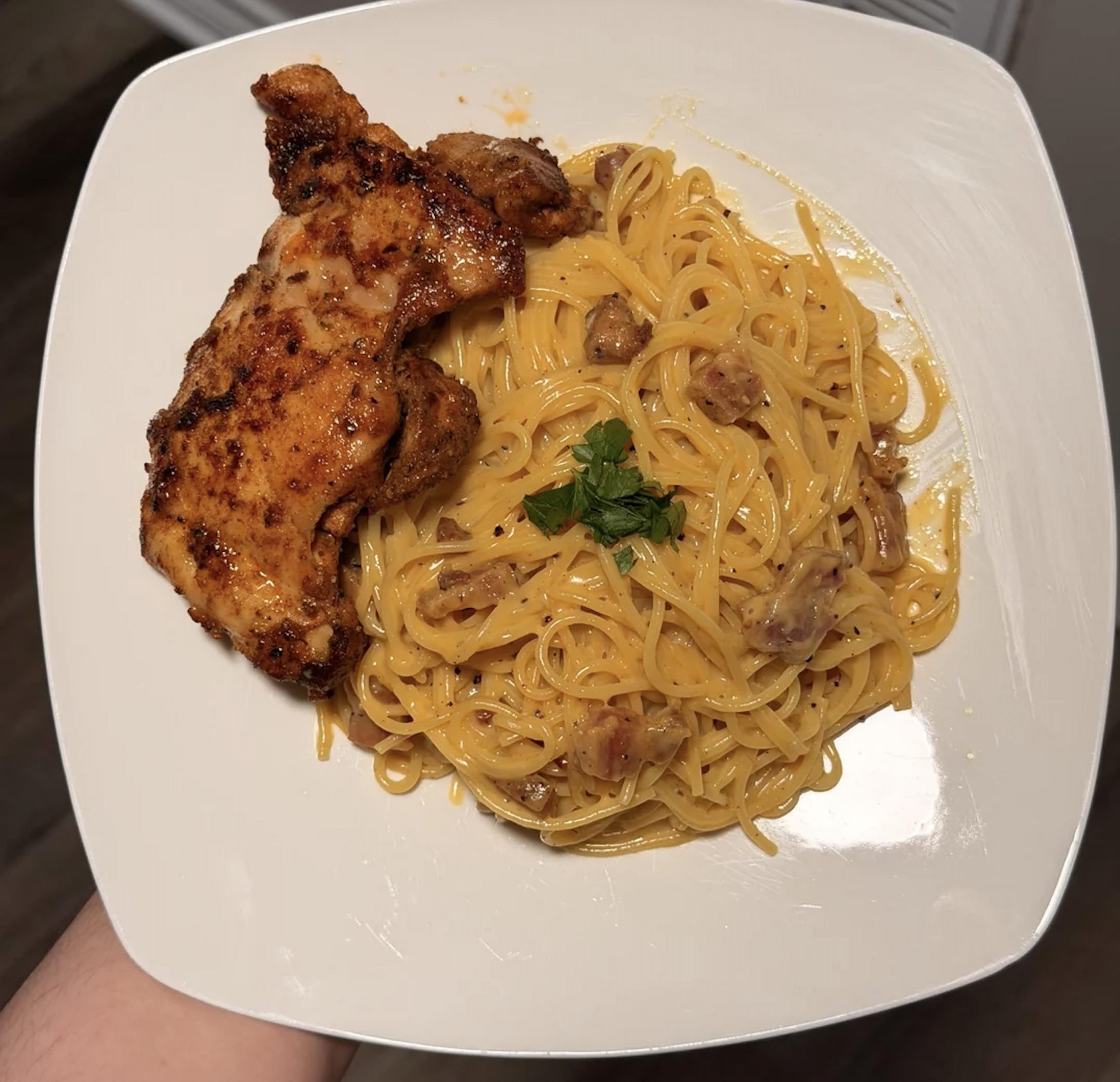 A white plate with creamy spaghetti carbonara topped with parsley, served alongside a piece of grilled, seasoned chicken.