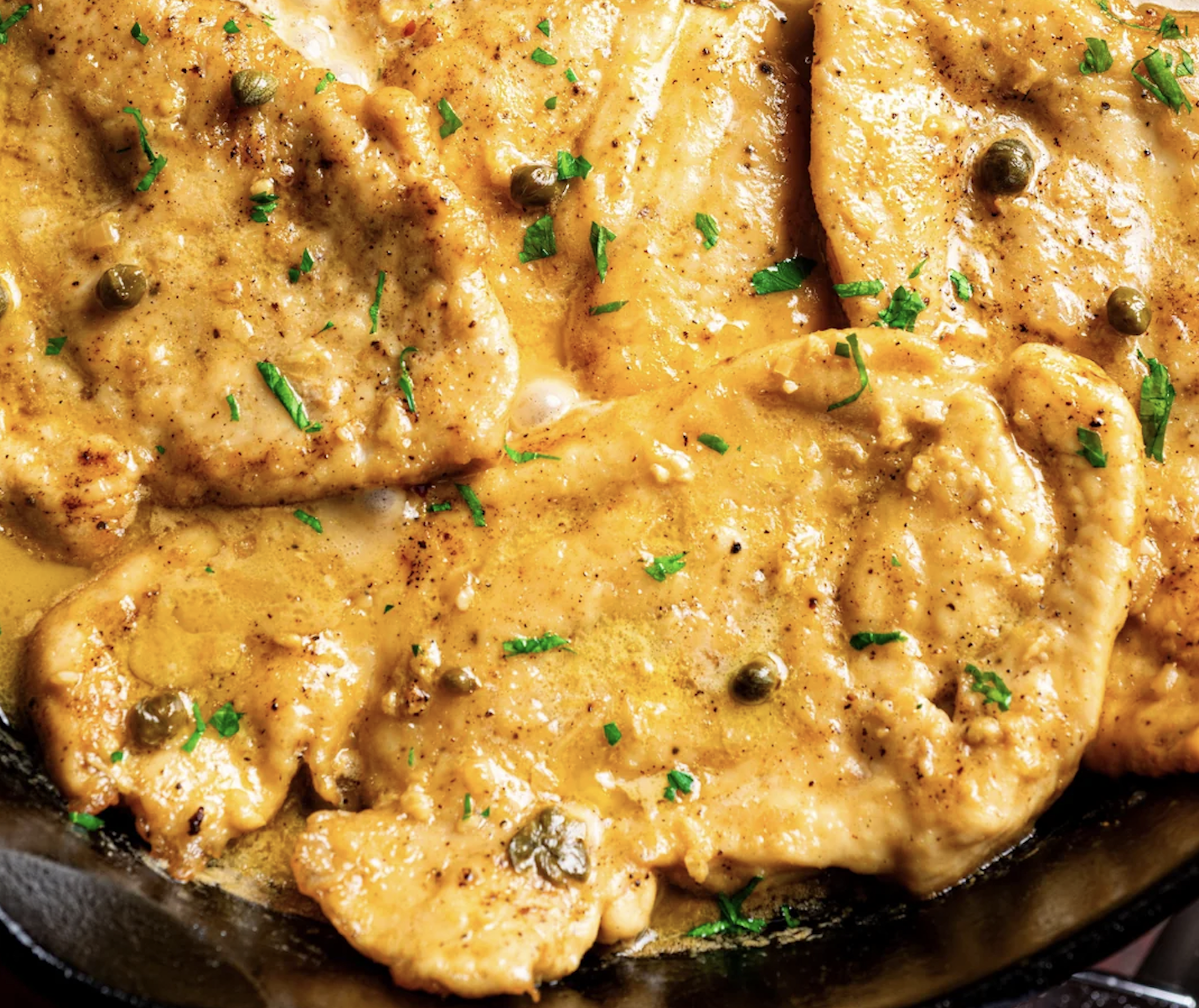 Golden-brown chicken cutlets in a lemony sauce, garnished with chopped parsley and capers, served in a skillet.