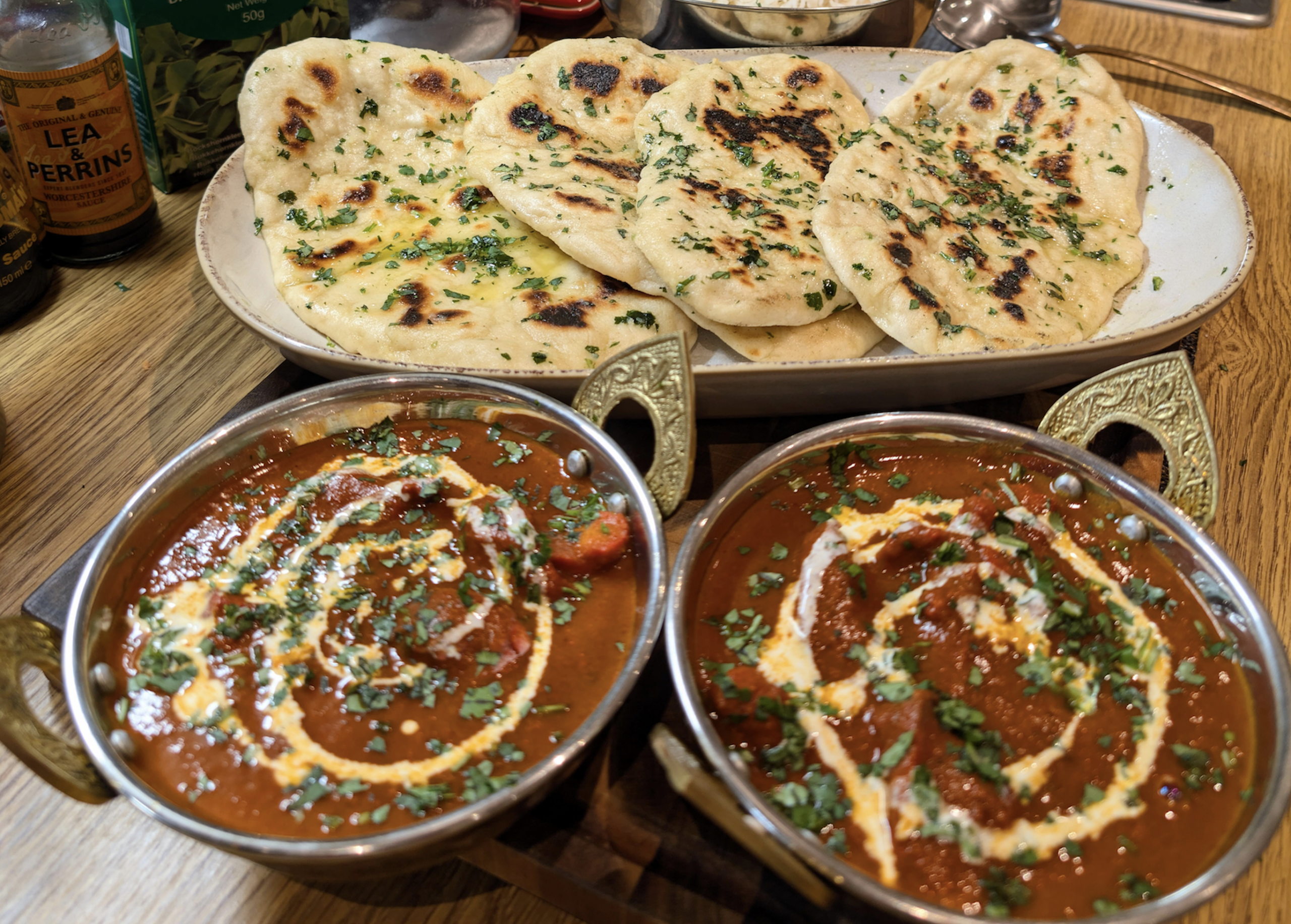 Two bowls of creamy curry garnished with herbs and cream sit in front of a plate of naan bread, all displayed on a wooden table with sauce bottles in the background.