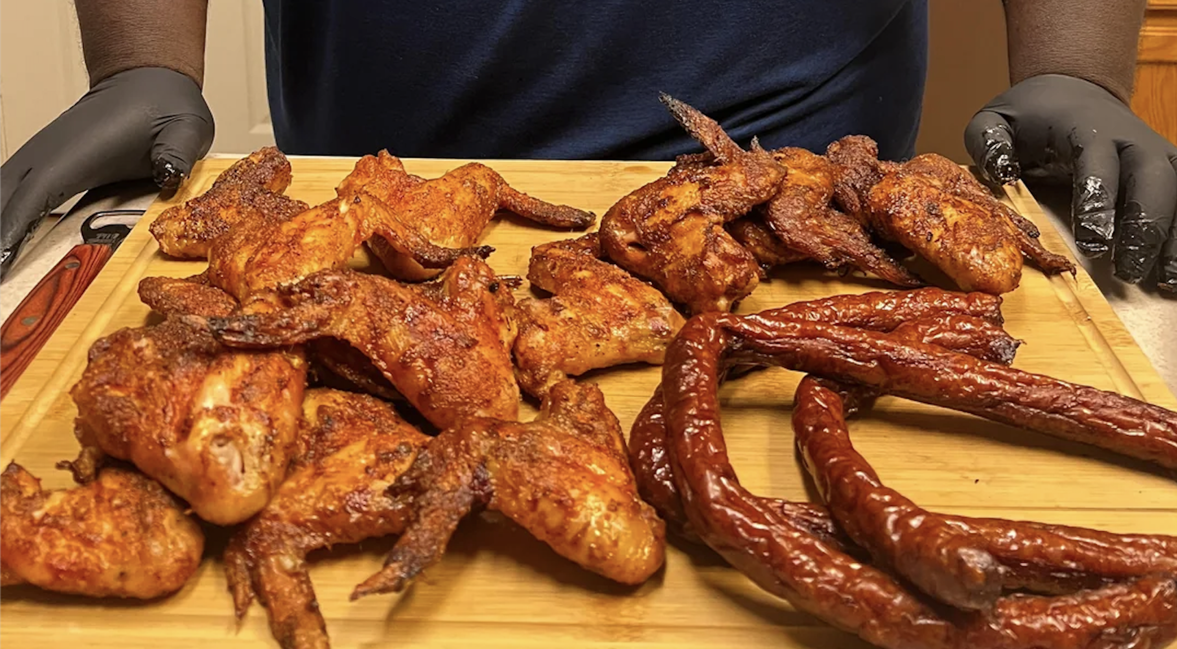 A person wearing black gloves presents a wooden board with seasoned, cooked chicken wings and smoked sausages.