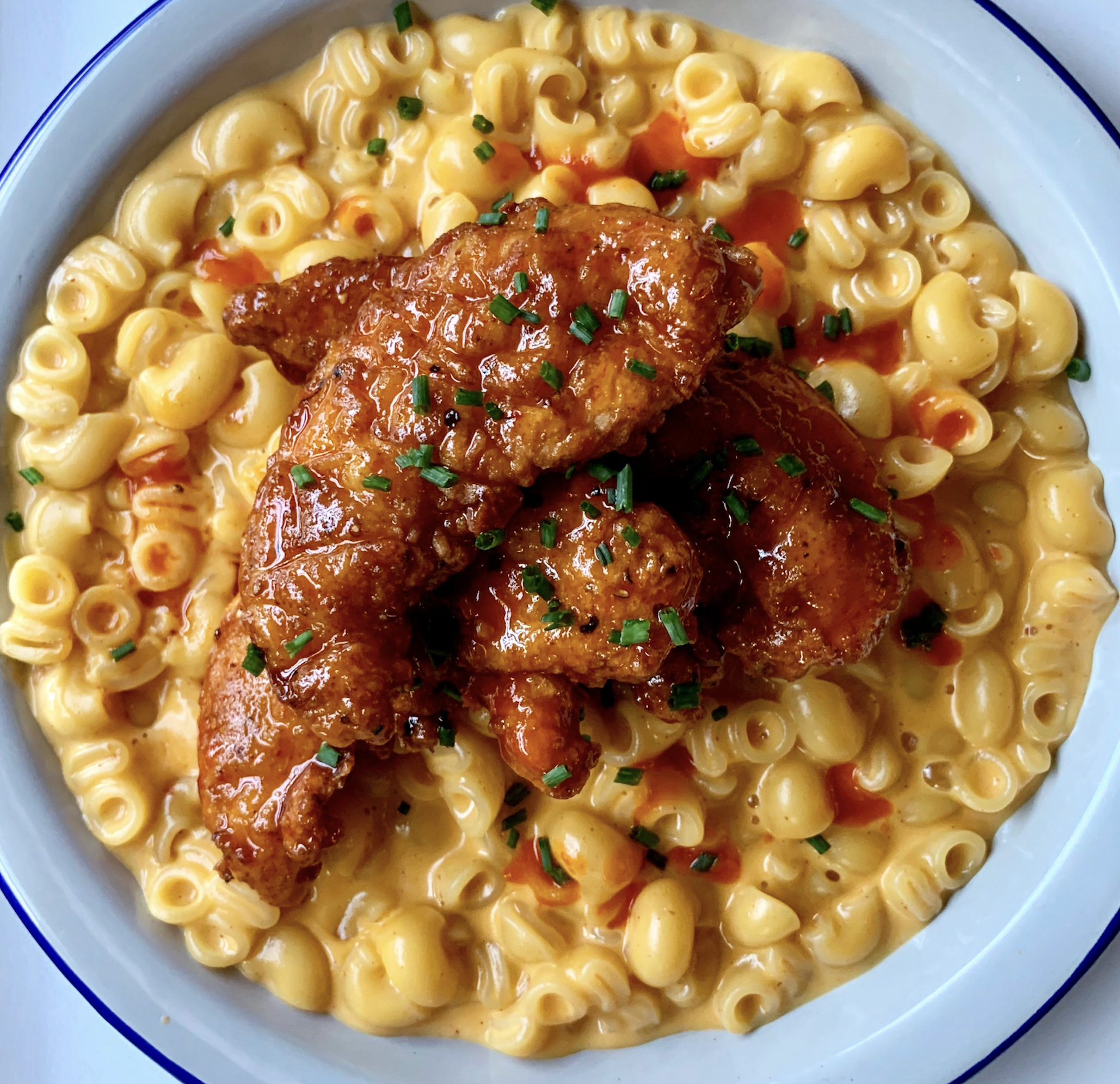 A bowl of creamy macaroni and cheese topped with crispy, sauced chicken tenders, garnished with chopped chives and a drizzle of hot sauce.