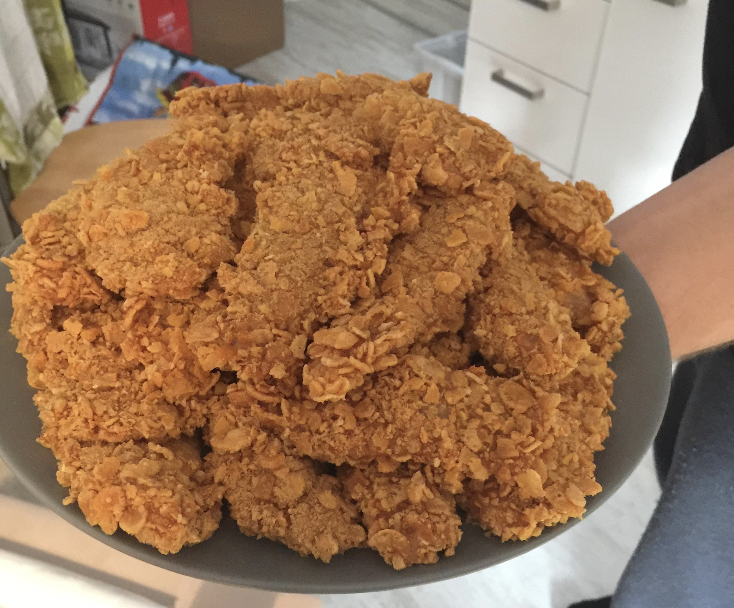 A gray plate piled high with crispy, golden-brown fried chicken strips is being held by a person in a kitchen.