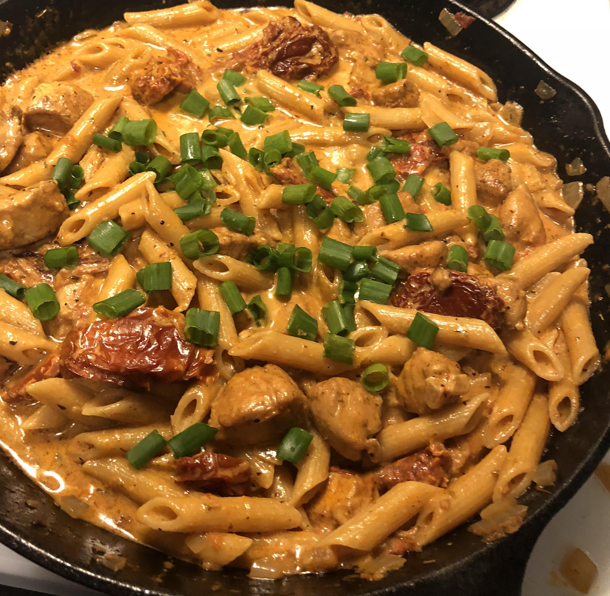 Creamy penne pasta with chunks of chicken, sun-dried tomatoes, and green onions served in a black skillet.