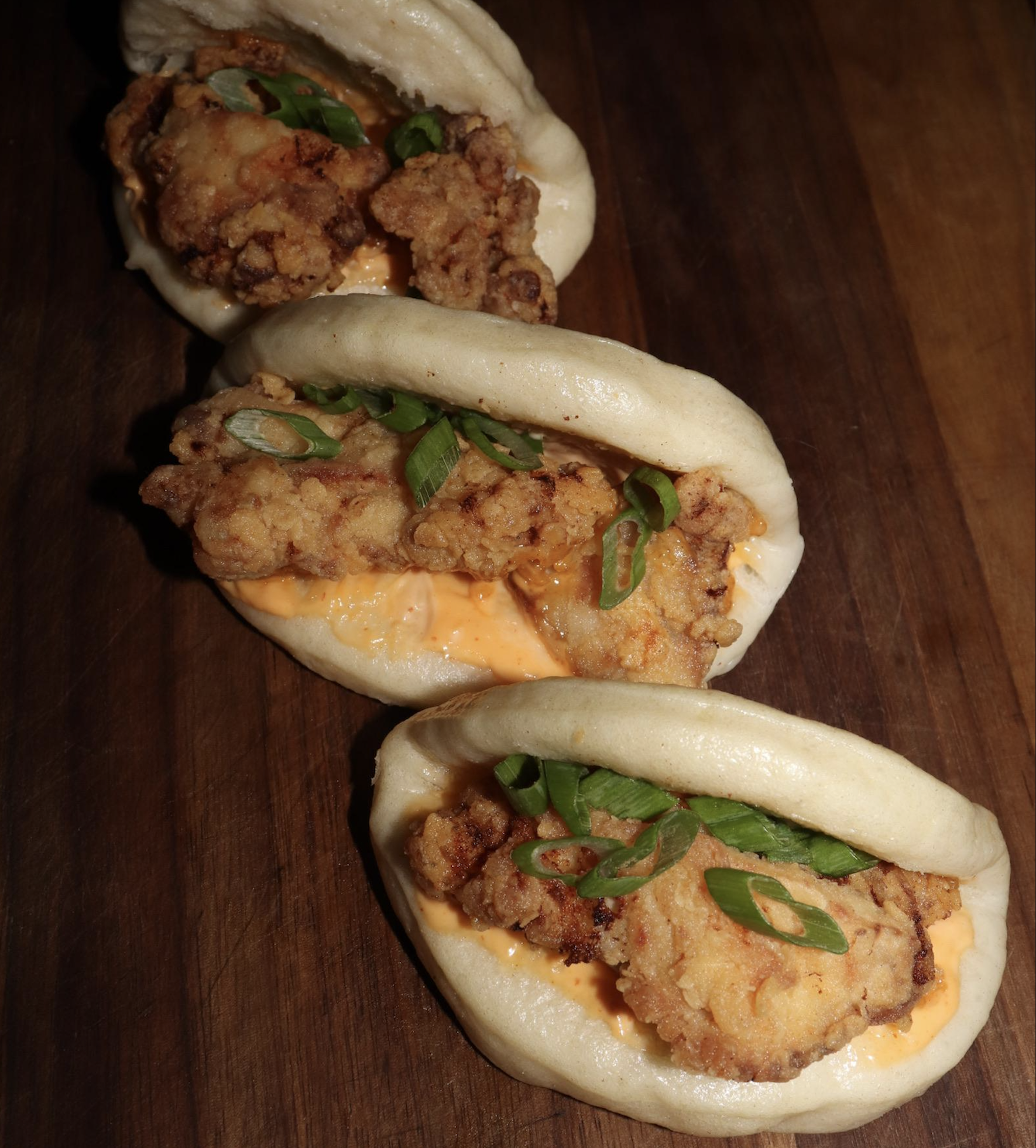 Three bao buns filled with crispy fried chicken, spicy orange sauce, and sliced green onions, arranged on a wooden surface.