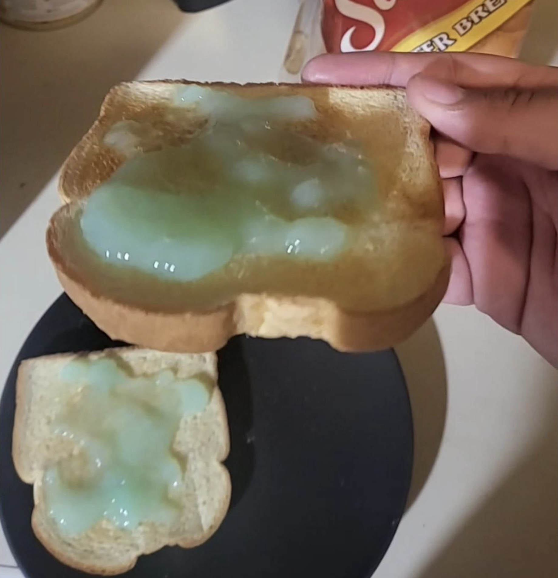 A hand holds a slice of toast topped with a greenish spread; another piece with the same spread is on a black plate below. A bread bag is partially visible in the background.