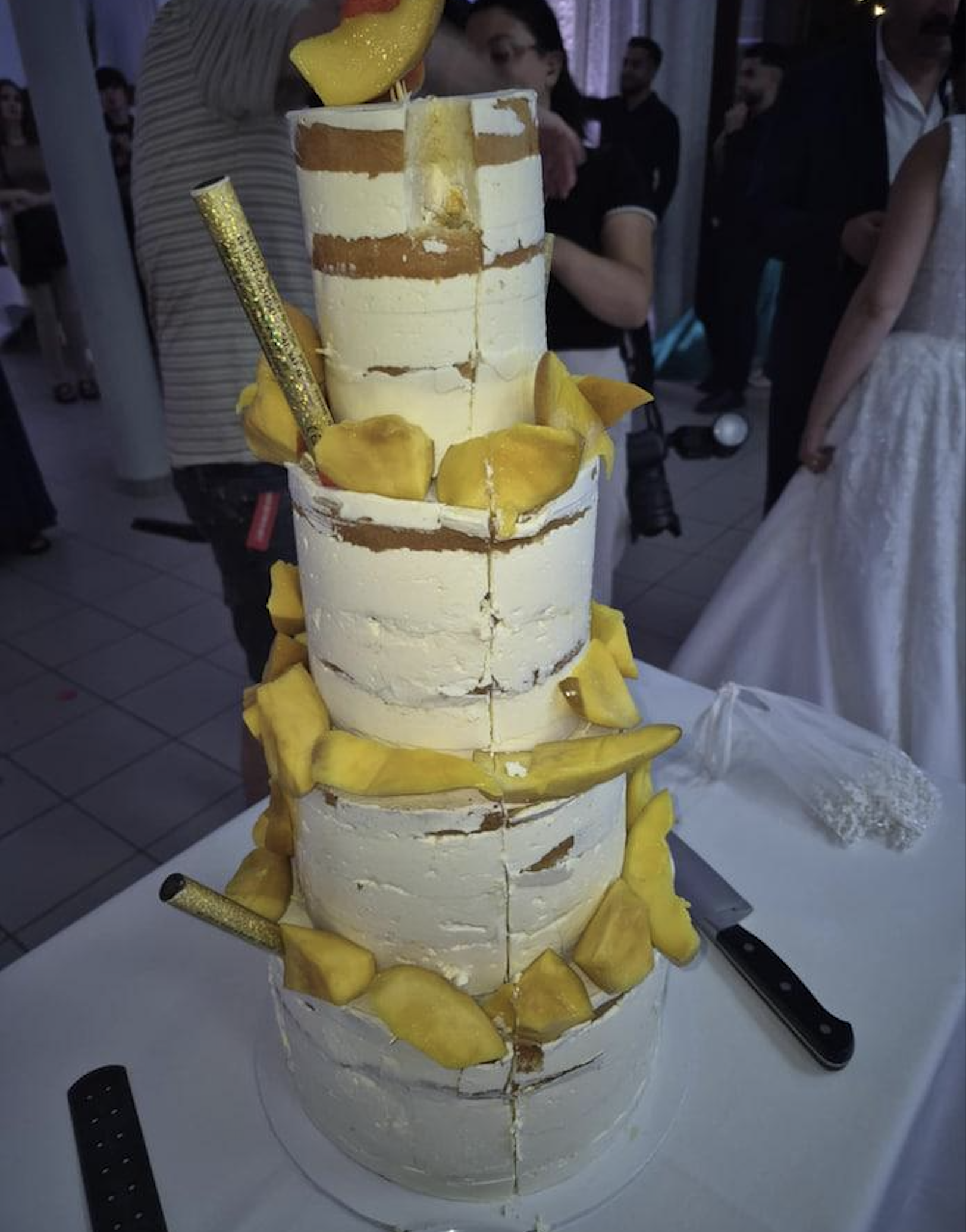 A four-tiered naked cake with white frosting and yellow fruit slices, possibly mango, is displayed on a table. People in formal attire are visible in the background.