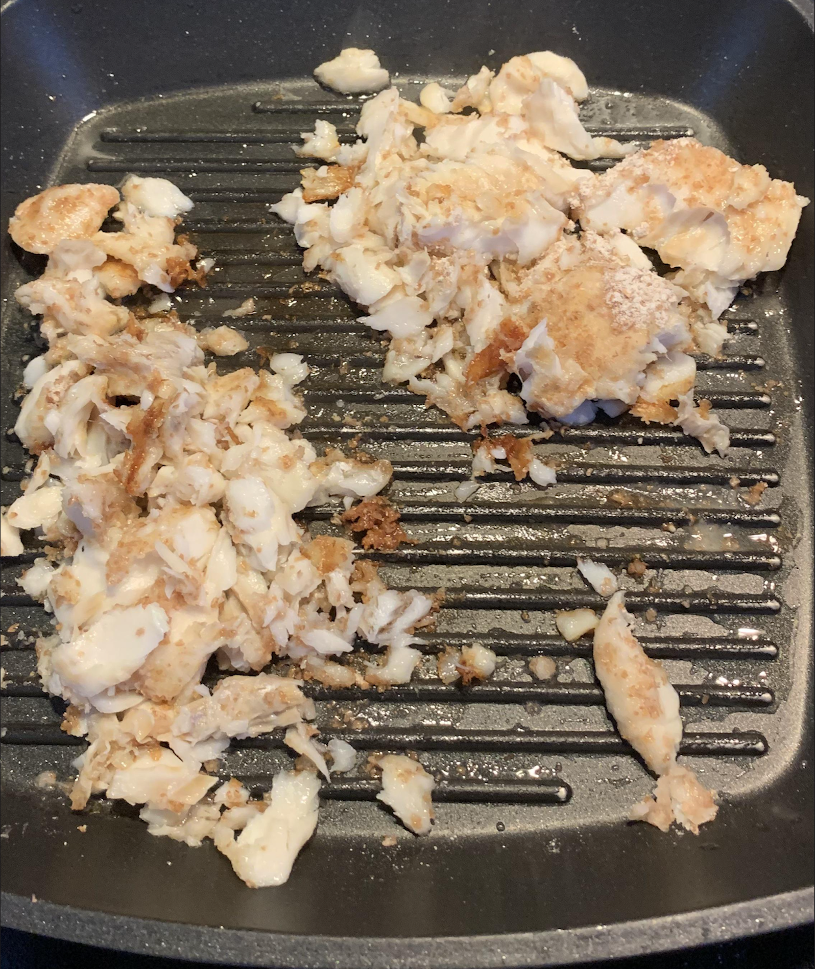 Cooked, broken pieces of white fish are scattered on a ridged grill pan. The fish appears flaky and slightly browned in spots, covering one side of the pan while the other side is mostly empty.