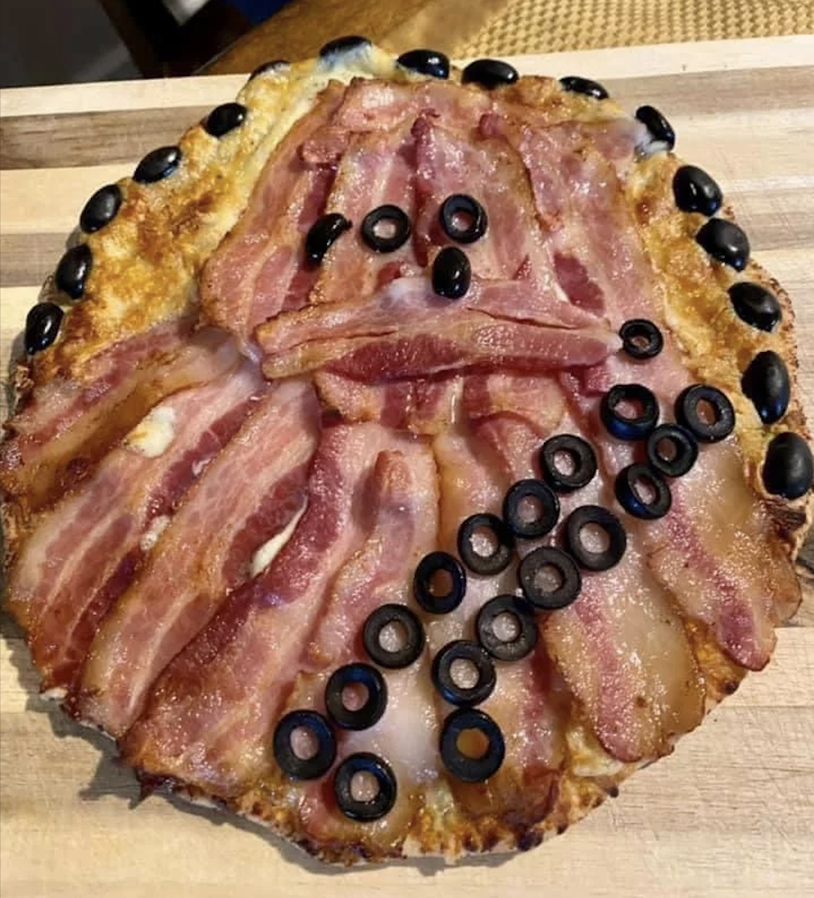 A pizza decorated to look like Chewbacca from Star Wars, using strips of bacon for fur and sliced black olives for eyes, nose, and a bandolier across the body.