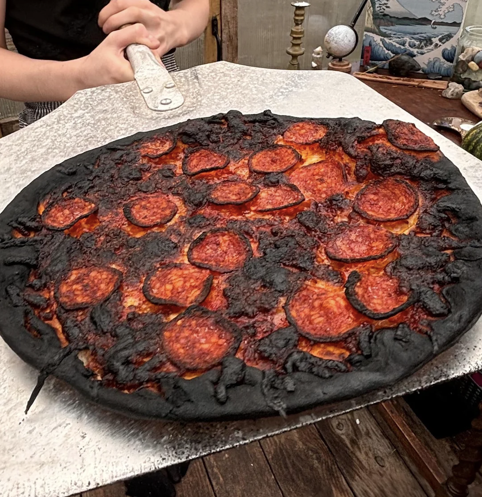 A person holds a pizza paddle with a heavily burnt pepperoni pizza, featuring blackened crust and melted cheese, on a rustic wooden table.