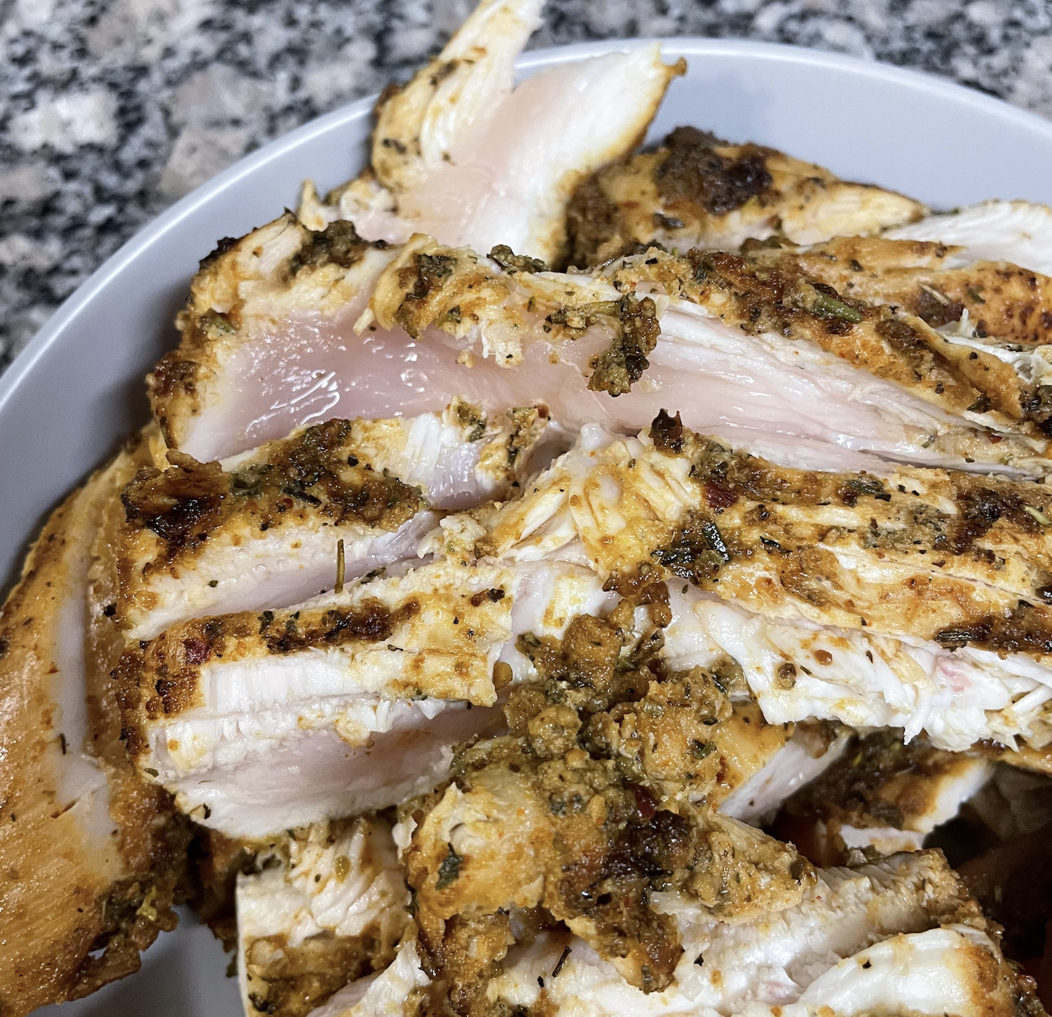 Sliced grilled chicken breast with visible pink, undercooked centers on a white plate, set on a granite countertop. The seasoning is visible on the surface of the meat.