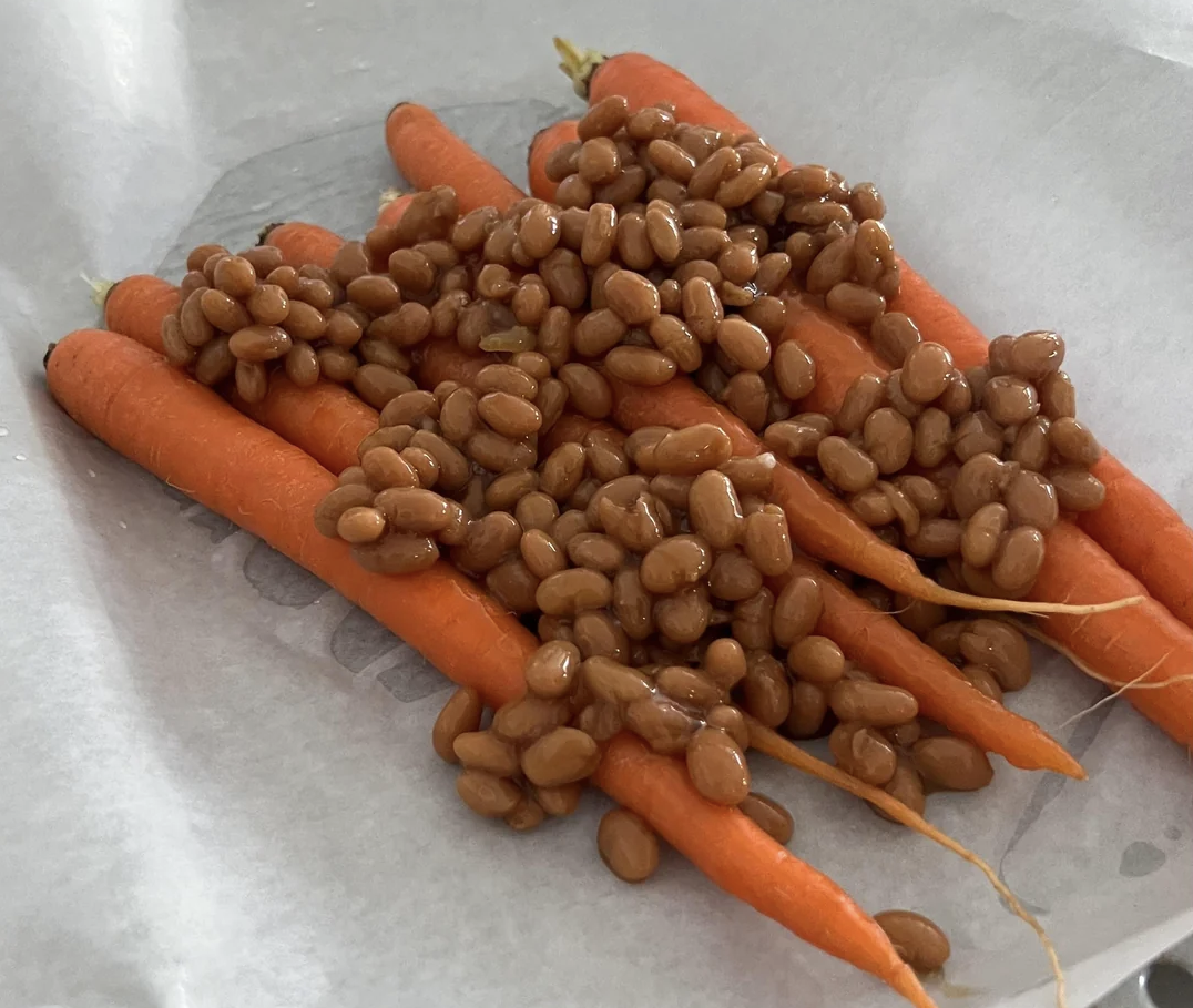 Whole baby carrots topped with baked beans are arranged on a sheet of parchment paper, ready for preparation or serving.