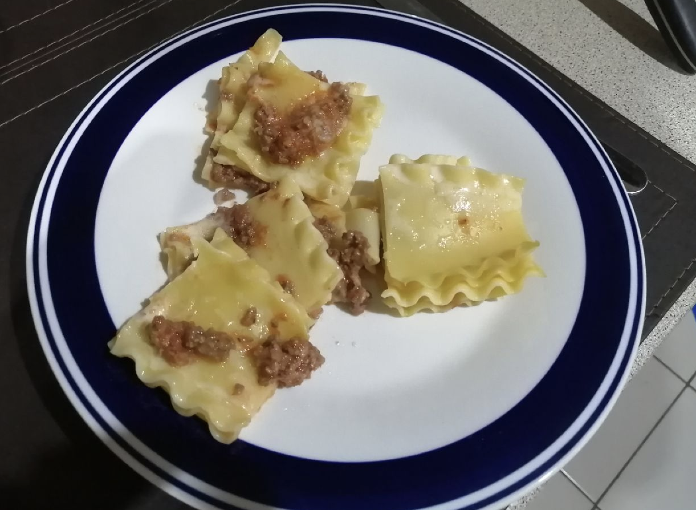 Three pieces of lasagna with visible meat sauce and cheese are served on a white plate with a blue rim, sitting on a counter.