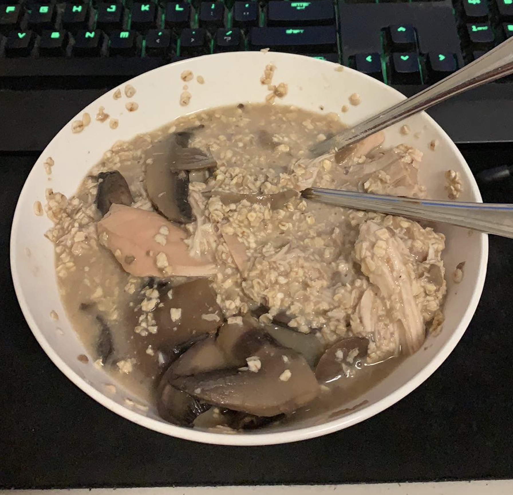 A bowl of oatmeal mixed with chunks of chicken and sliced mushrooms, with metal chopsticks resting inside. The bowl is placed on a black surface in front of a computer keyboard.