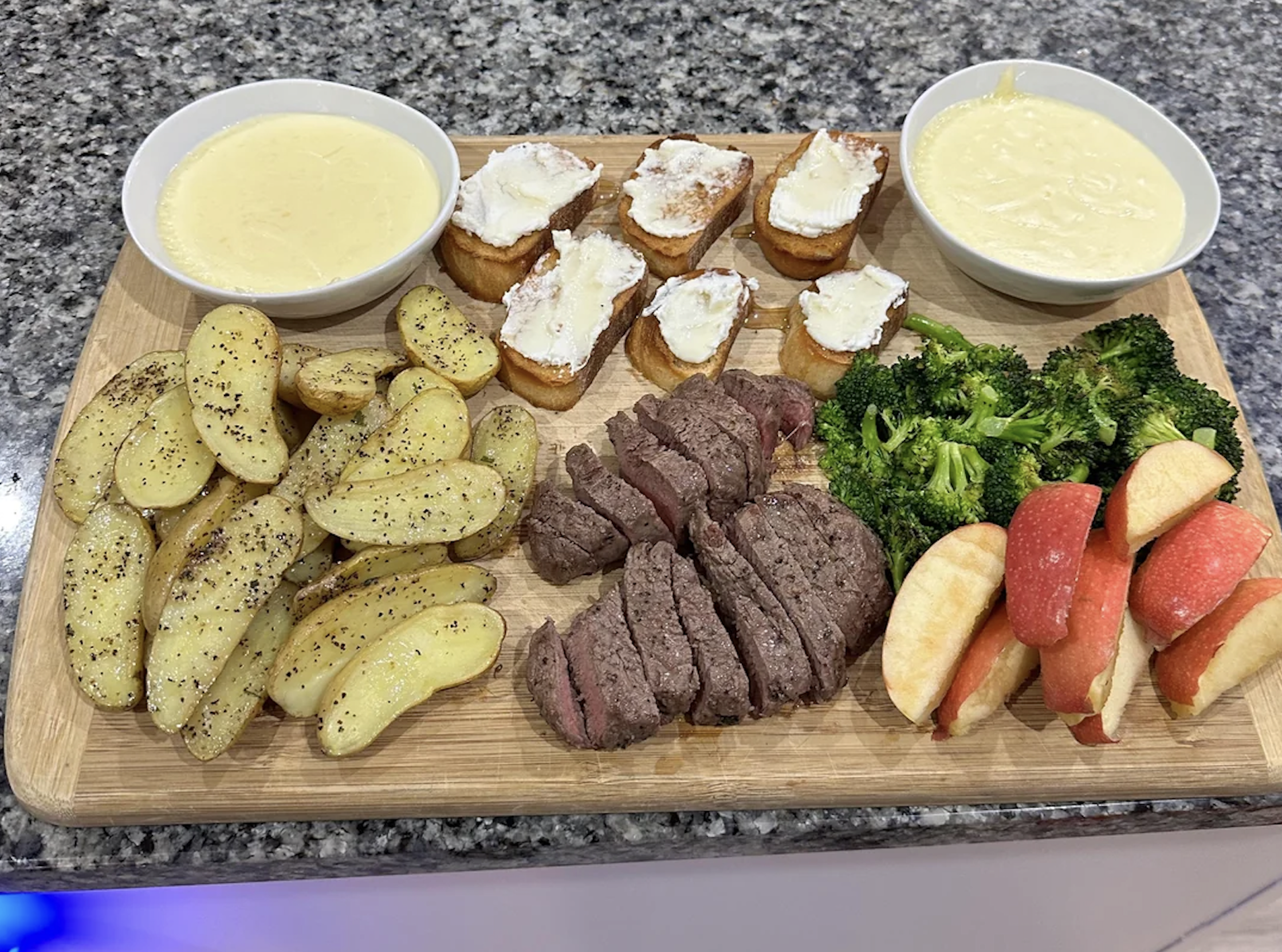A wooden board with sliced steak, roasted potato wedges, broccoli, apple slices, toasted bread with cheese, and two bowls of cheese fondue on a granite countertop.