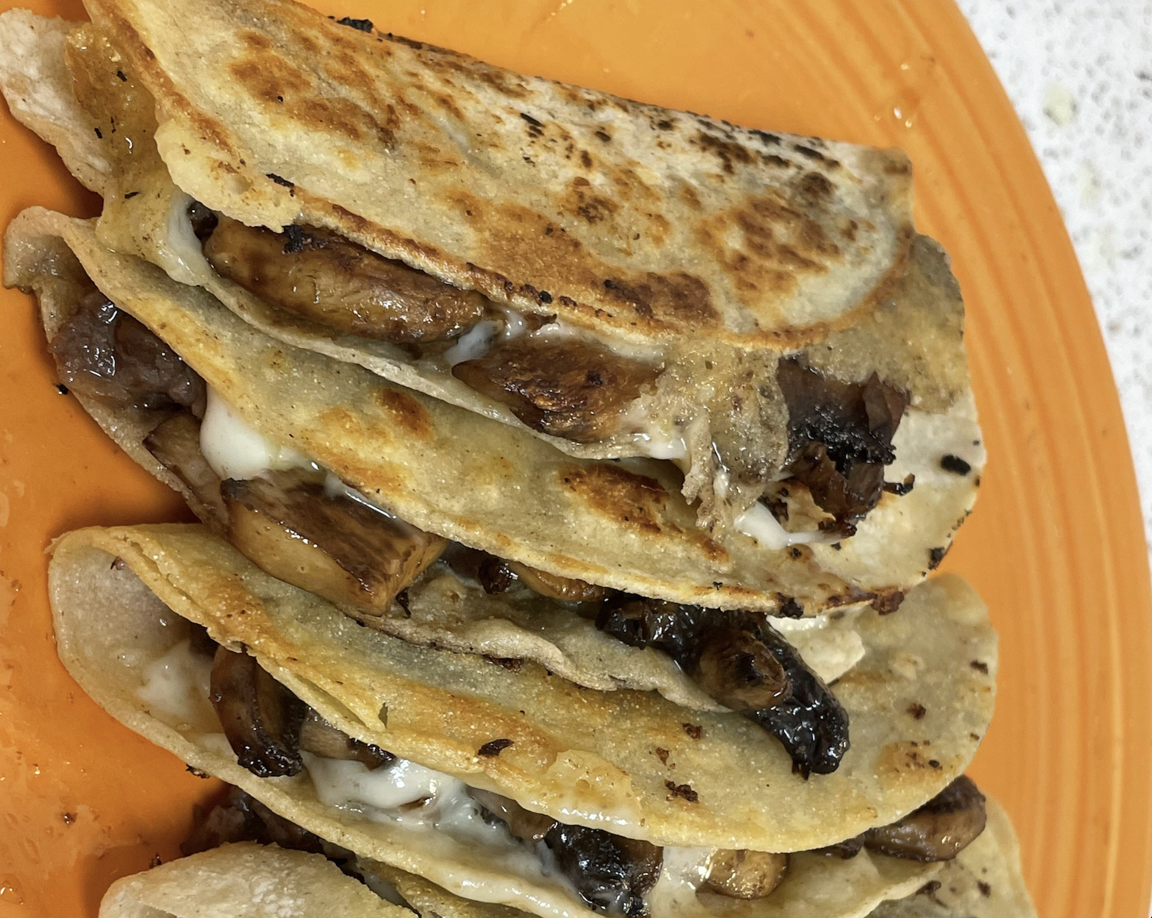 Three folded tacos filled with melted cheese and grilled mushrooms, stacked on an orange plate. The tortillas appear lightly toasted with golden brown spots.