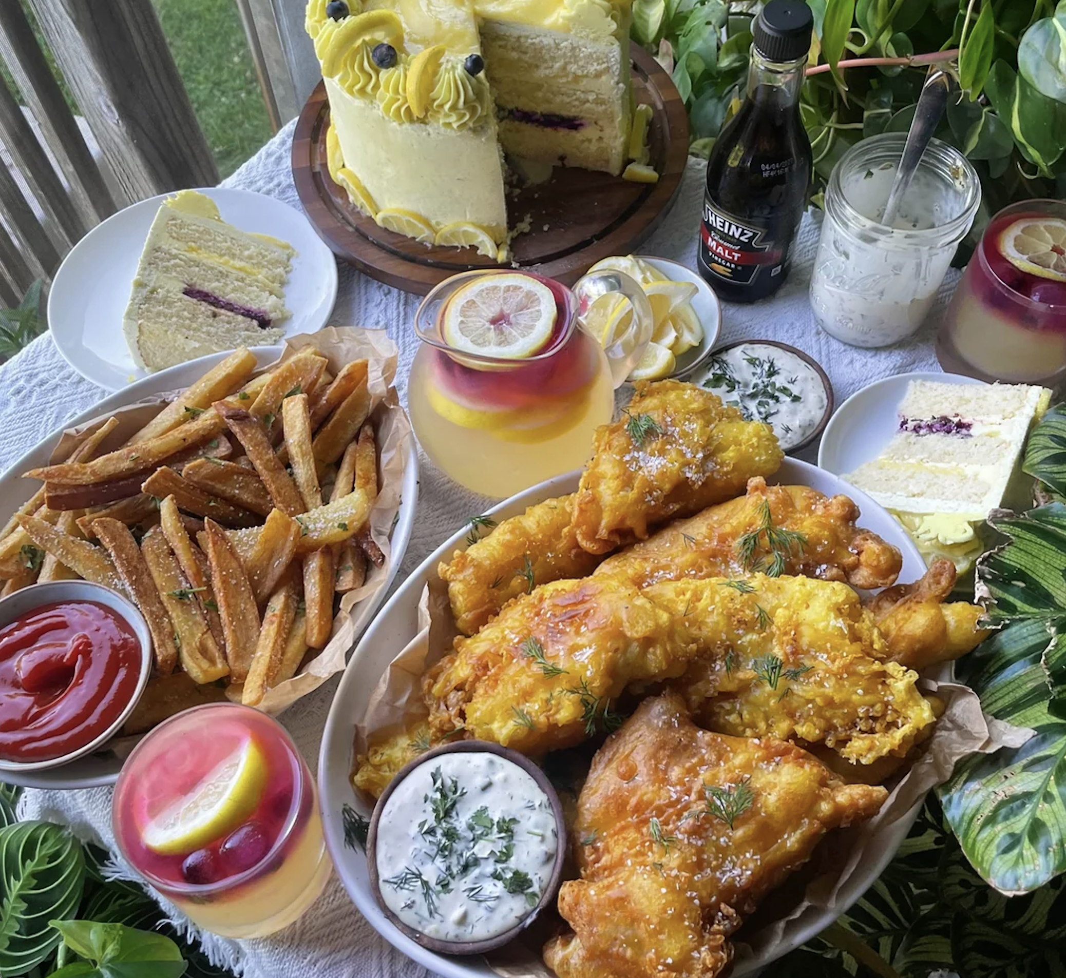 A table with fried fish, French fries, lemon drinks, ketchup, tartar sauce, slices of lemon layer cake, and a bottle of malt vinegar, arranged with leafy greenery in the background.