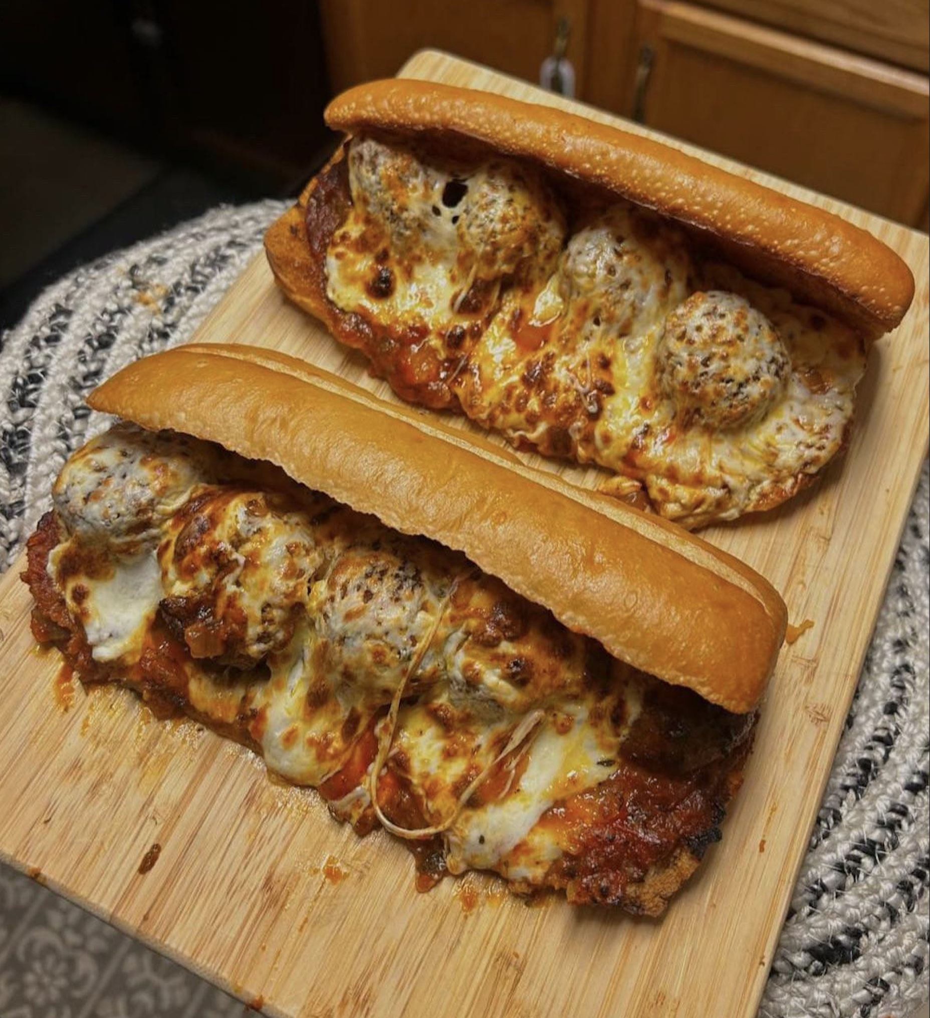 Two meatball subs with melted cheese and marinara sauce on toasted buns are placed on a wooden cutting board.
