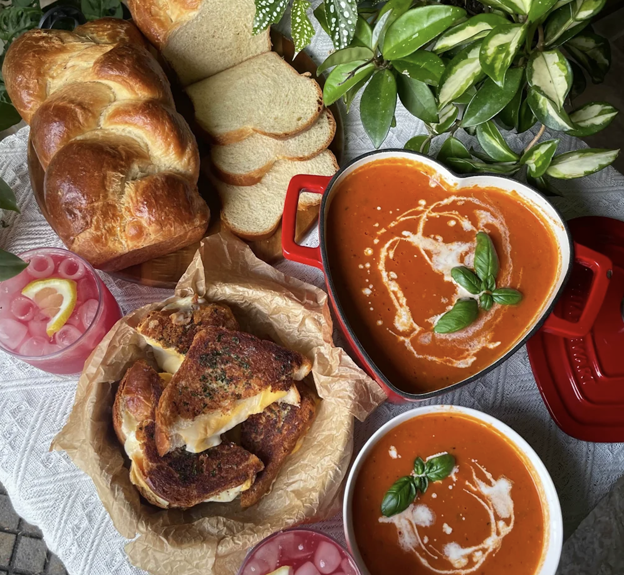 Heart-shaped bowls of tomato soup with cream and basil, grilled cheese sandwiches, braided bread, sliced white bread, and two pink drinks with lemon, arranged on a table with green plants.