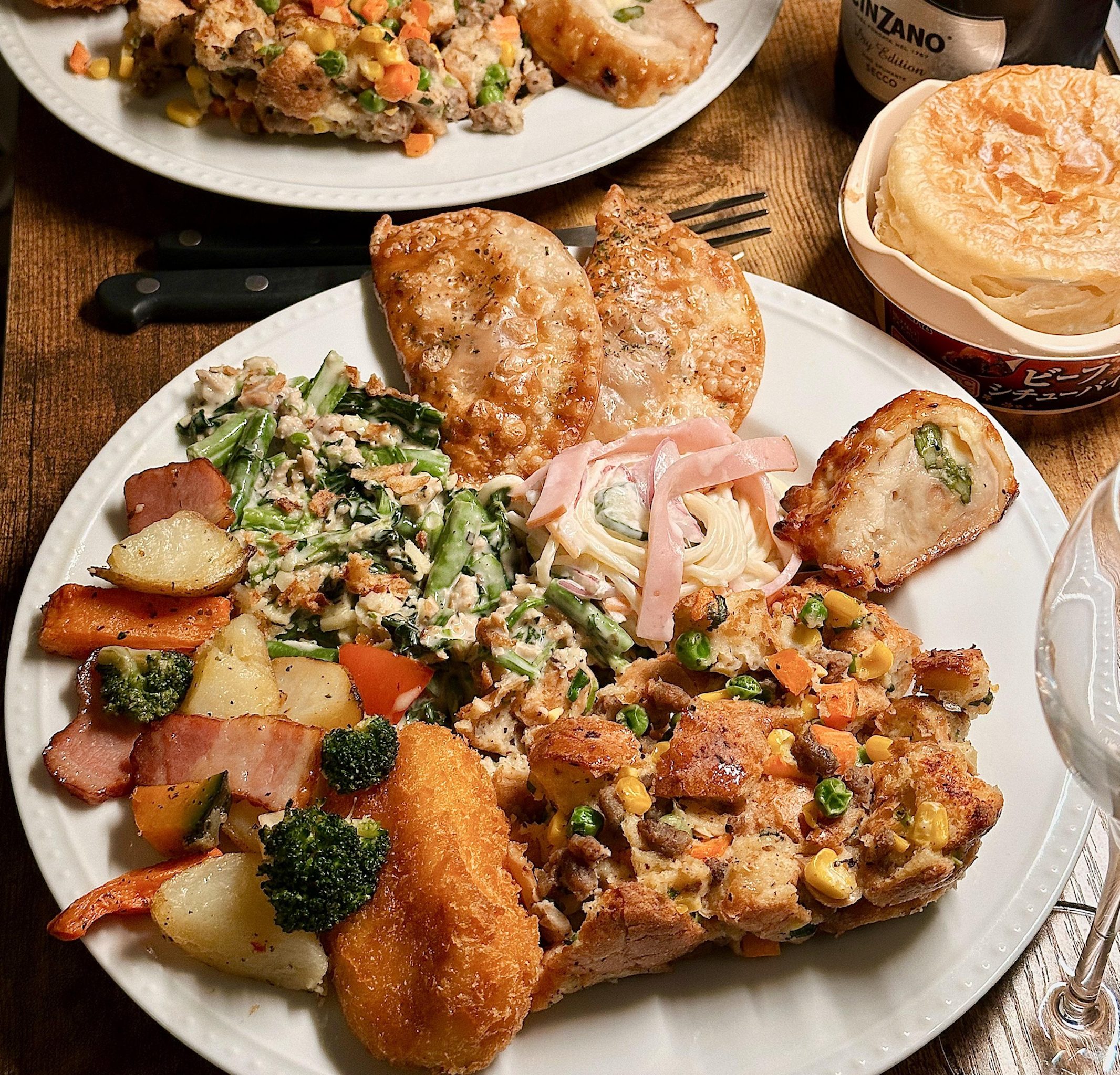 A dinner plate with roasted potatoes, broccoli, breaded cutlet, salad, pasta with ham, mixed vegetables, and two slices of meat. Another plate and a drink are partially visible in the background.