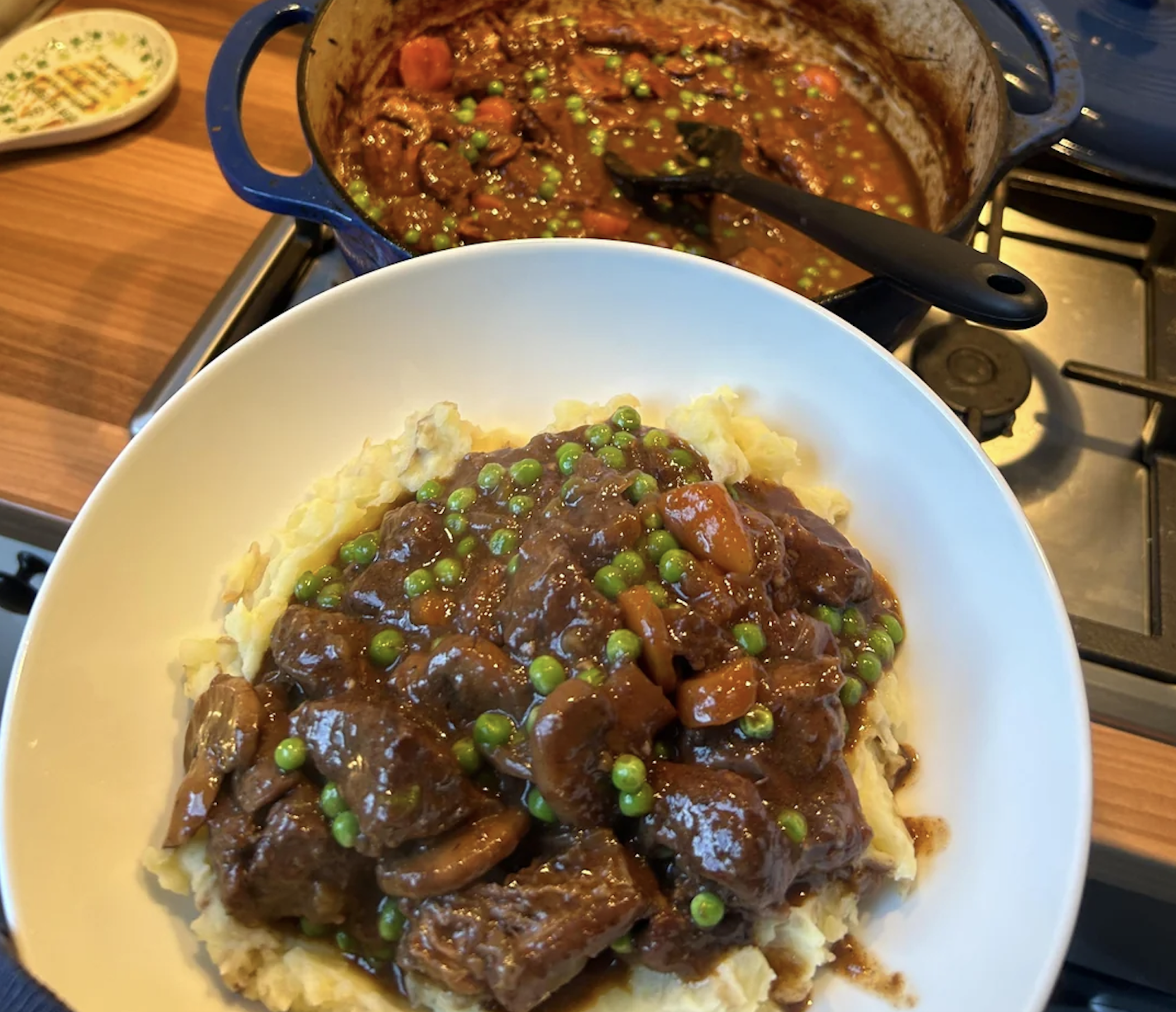 A white bowl filled with creamy mashed potatoes topped with a rich, dark beef stew containing carrots, mushrooms, and peas. A blue Dutch oven containing more of the same stew sits on the stovetop in the background.