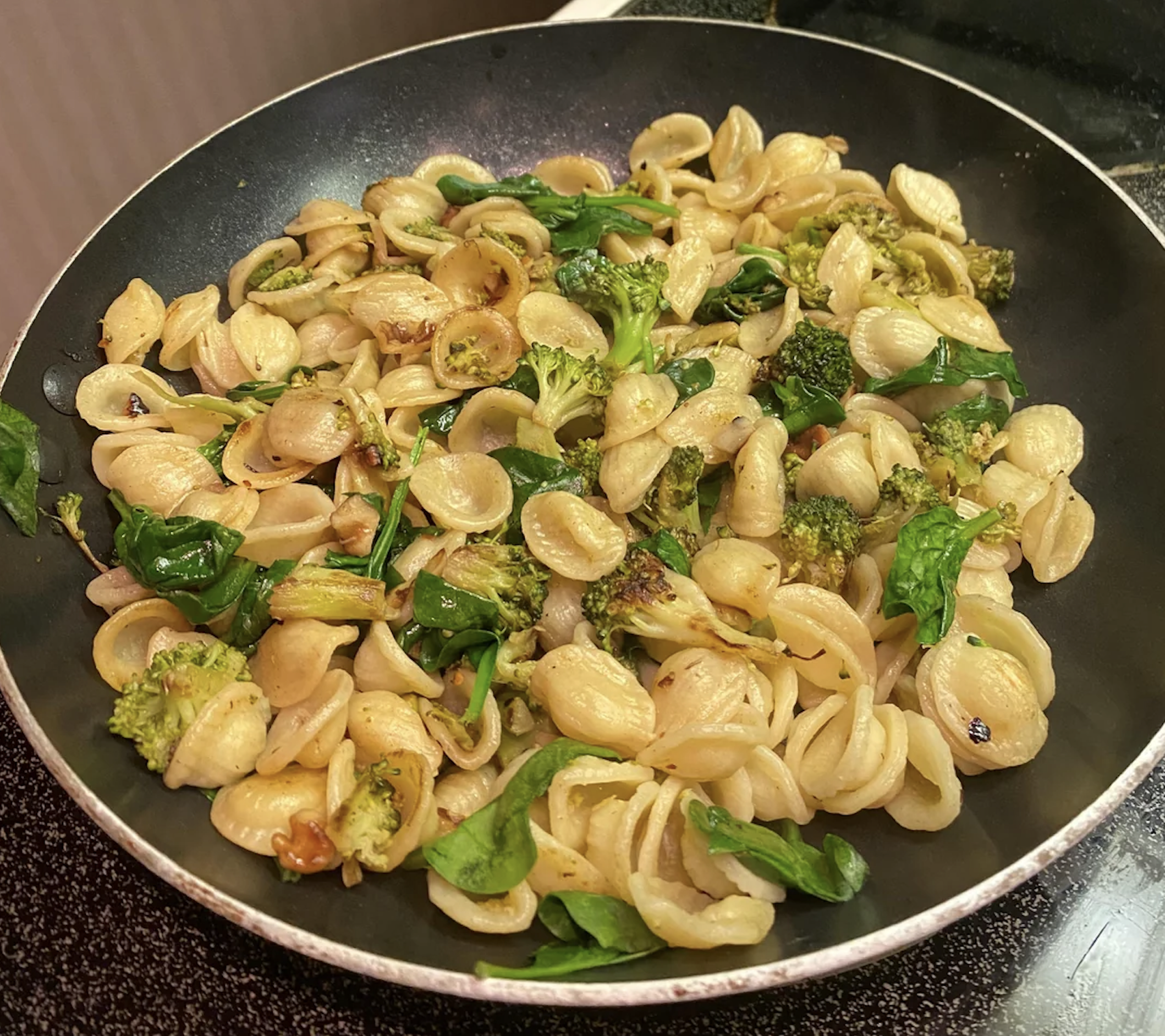 Orecchiette pasta in a black pan, cooked with broccoli, spinach, and light seasoning, sitting on a stovetop.