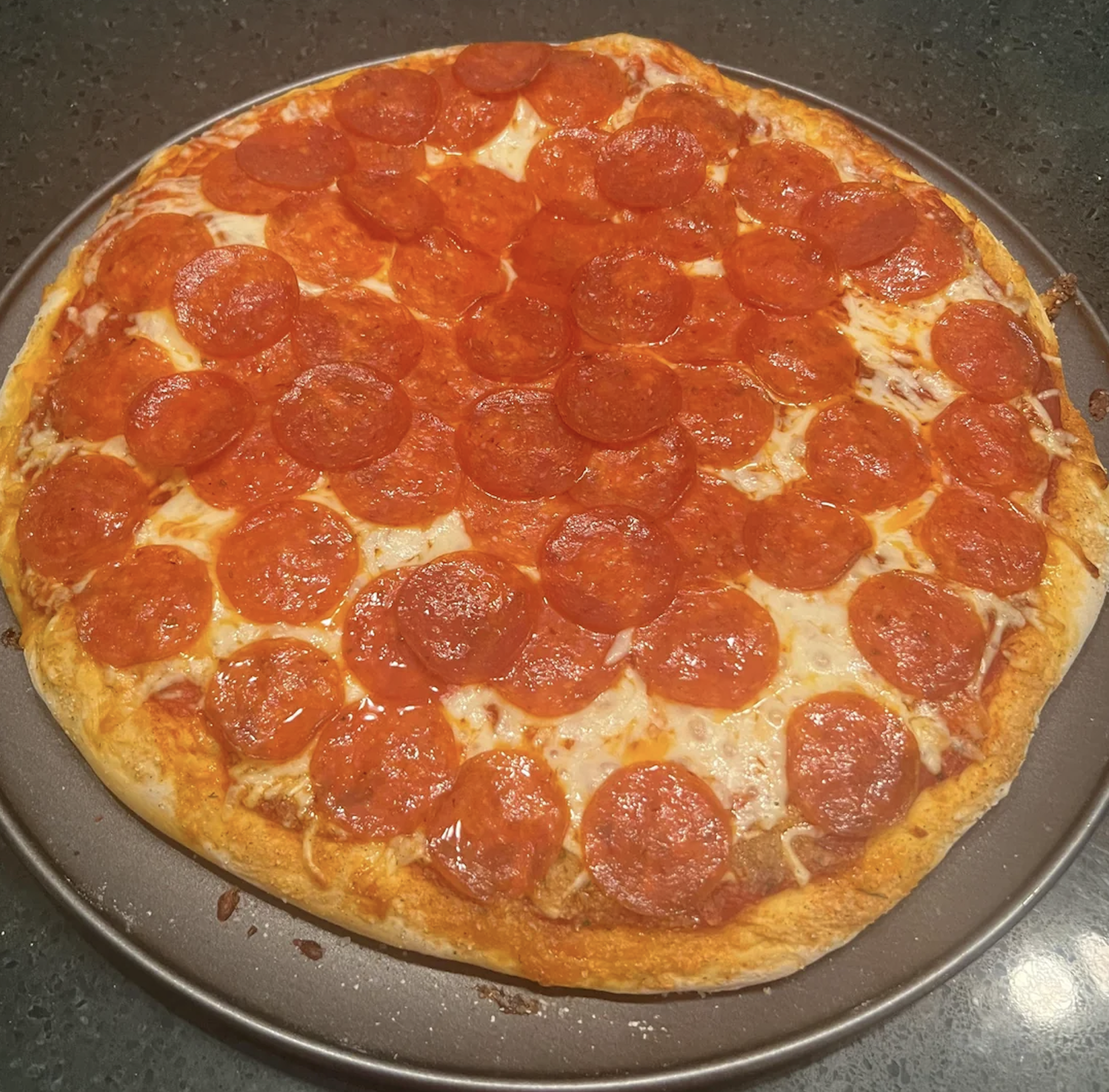 A freshly baked pepperoni pizza with melted cheese and a golden crust sits on a round baking tray on a dark countertop.