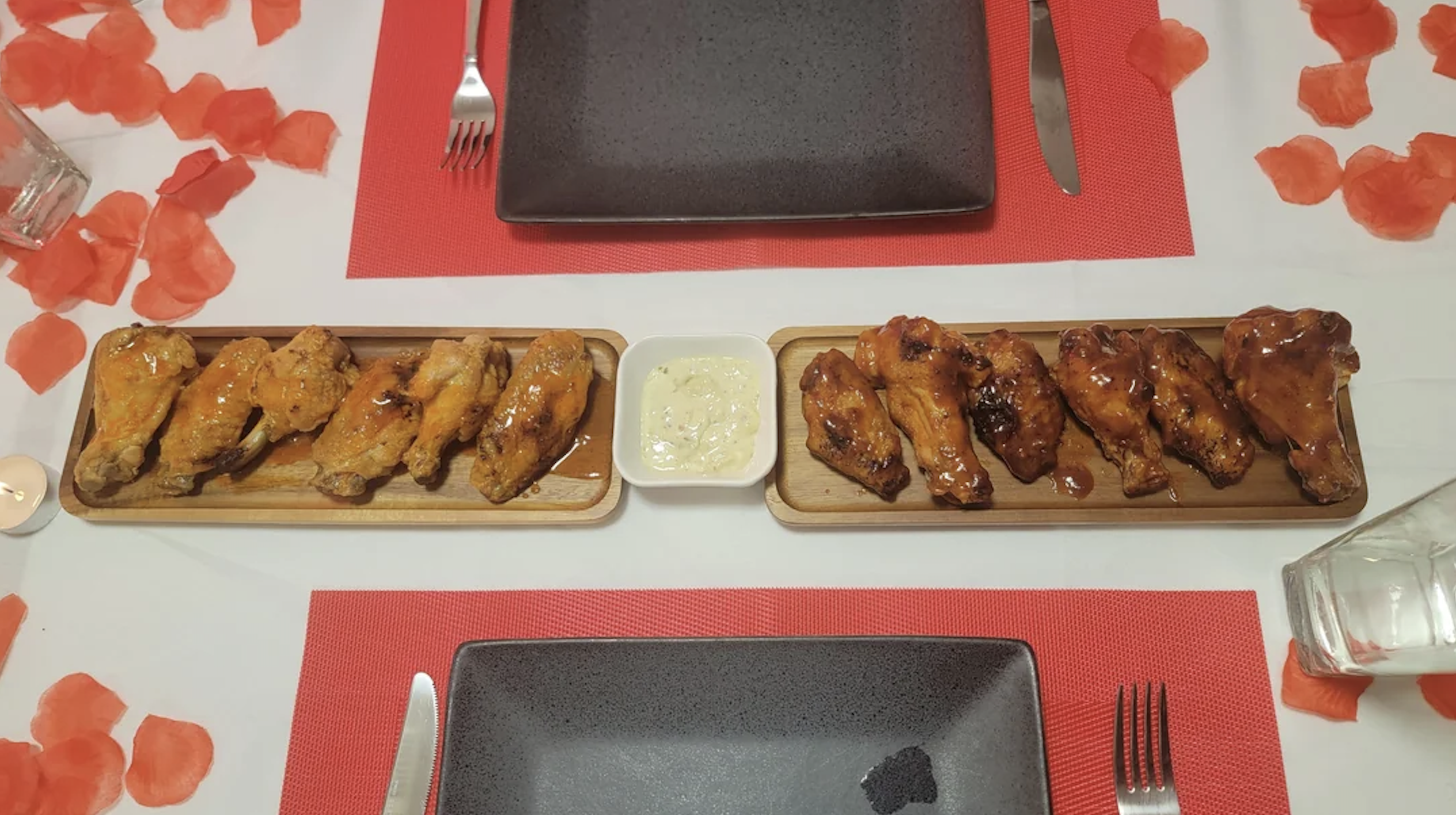 Two trays of chicken wings with sauce sit on a table set for two, with black plates, red placemats, silverware, and scattered red petals creating a romantic atmosphere.